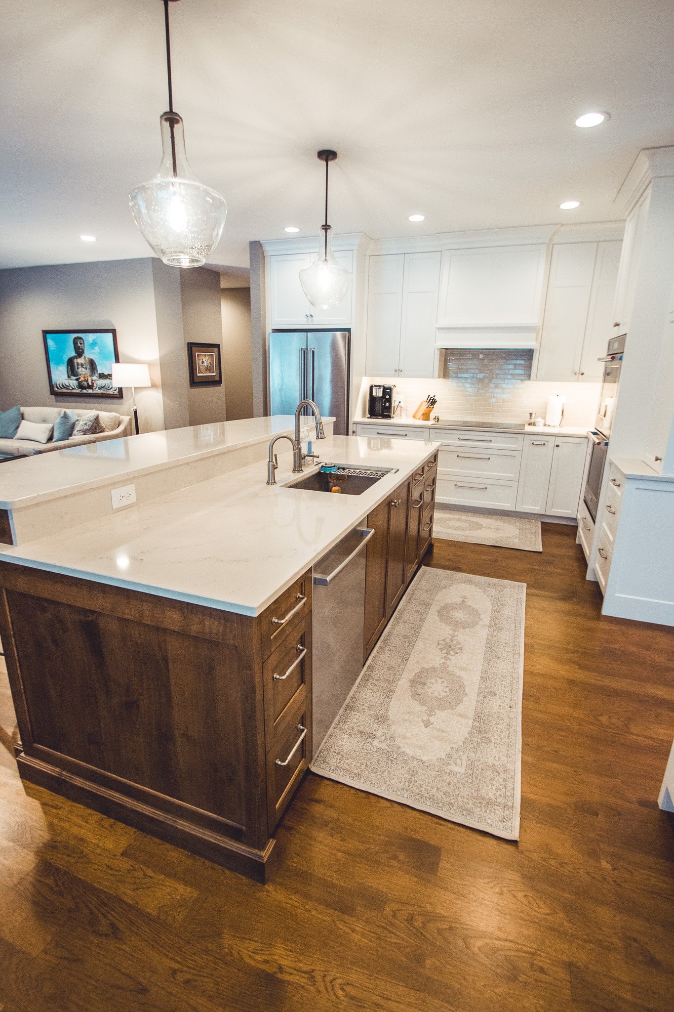 Modern kitchen remodel in Chanhassen with white cabinets, a large island with a wood base and white countertop, stainless steel appliances, pendant lights, and a decorative rug on wooden flooring.