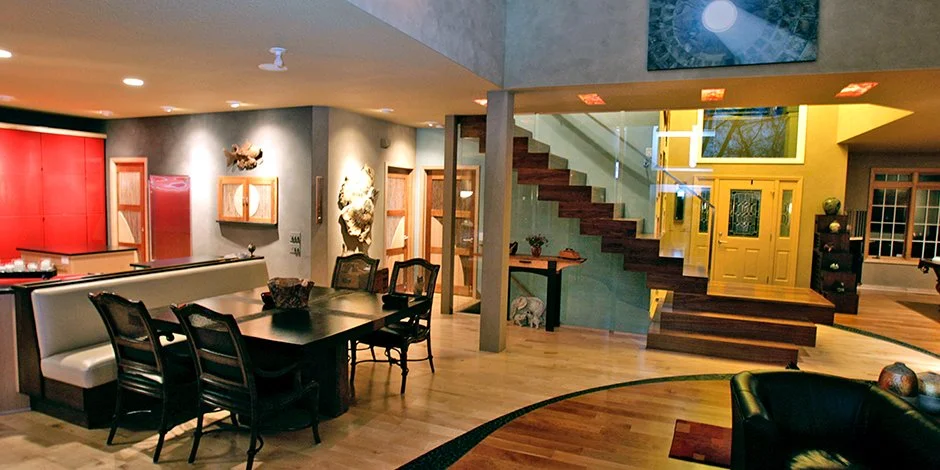 Modern open-concept living space  remodel in Excelsior with wooden flooring, featuring a dining area with a dark wood table and chairs, a brightly lit kitchen with red cabinetry, and a staircase with glass railing leading to the upper floor. 