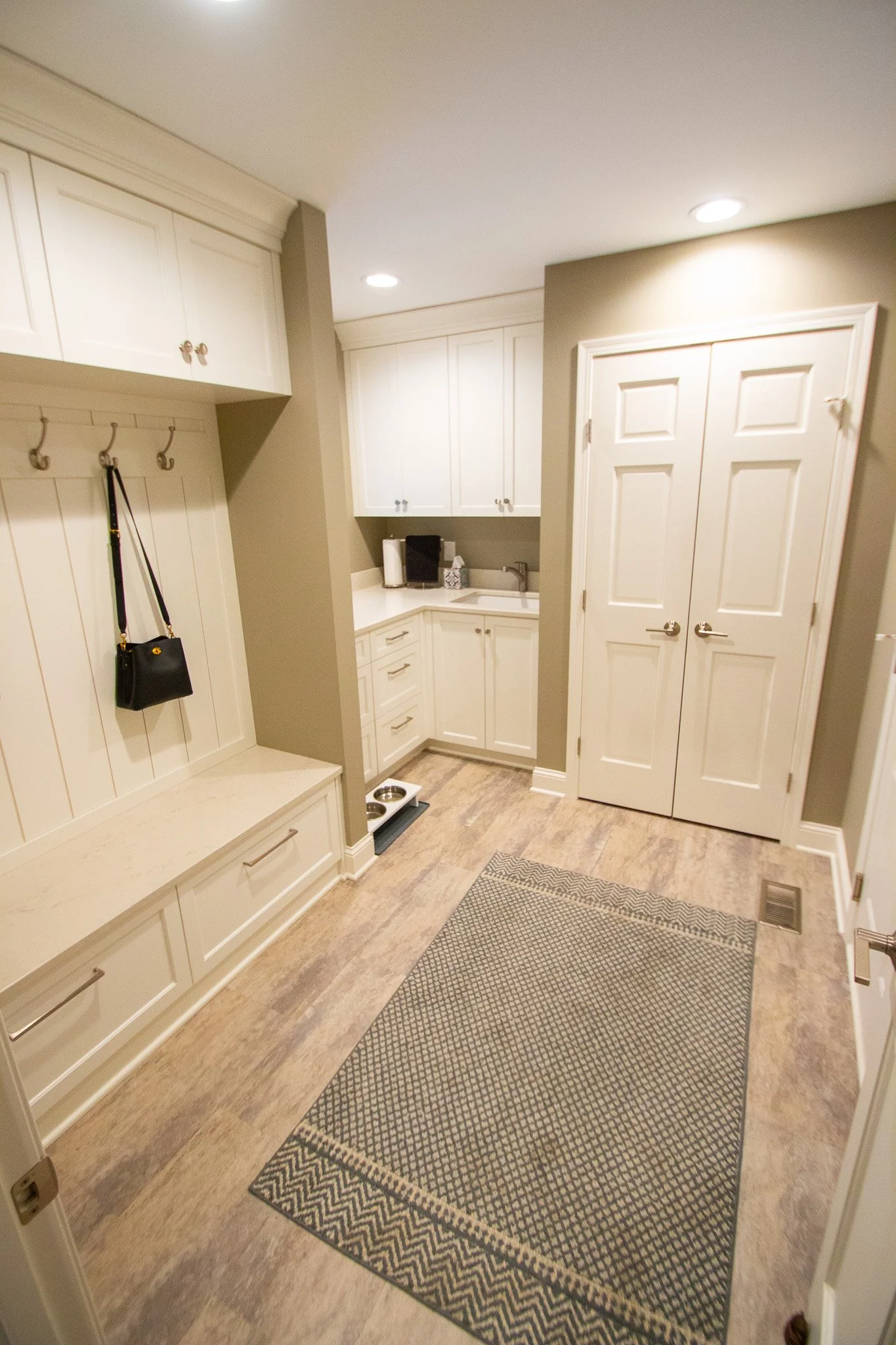Modern laundry room remodel in Eden Prairie with beige walls, white cabinets, and wood flooring. Includes a bench with hooks, a rug, double closet doors, and a countertop with a sink, paper towels, and storage cabinets.