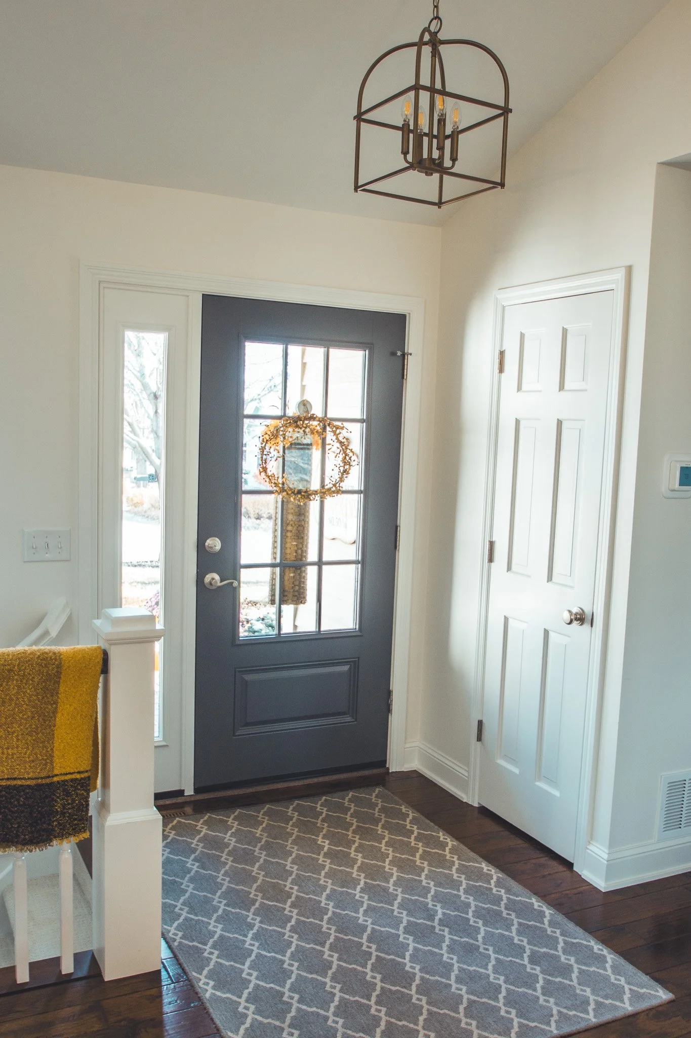 Interior entryway remodel in Minnetonka with a dark front door, adorned with a wreath. A patterned rug covers the wooden floor, and a yellow throw blanket is draped over the staircase railing. A modern pendant light hangs from the ceiling.