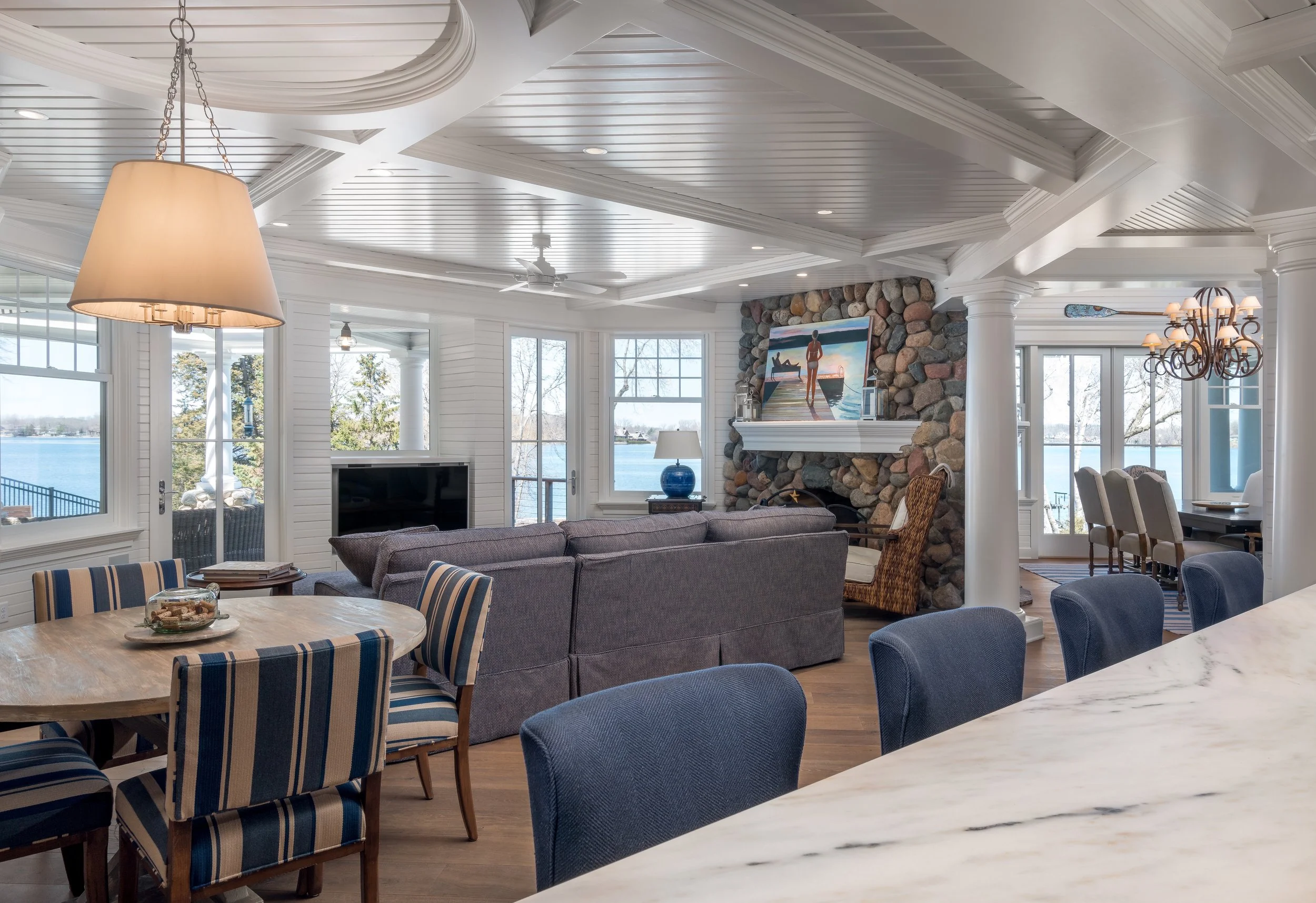 Luxurious living room remodel in Chanhassen with lake view, featuring a stone fireplace, artworks, dining area, striped chairs, and modern lighting.