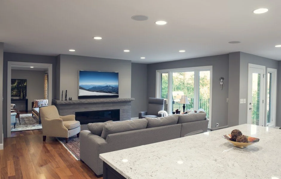 Modern  remodel in Excelsior with an open living room with gray sectional sofa, flat screen TV above fireplace, armchairs, and large windows providing natural light. Features hardwood floors and a view into the adjacent room.