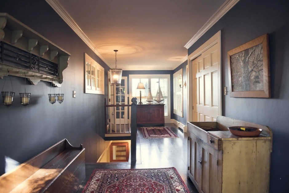 Traditional hallway remodel in Minnetonka with dark blue walls, wooden furniture, framed artwork, and decorative sconces. Features include a staircase, rug, and a window letting in natural light.