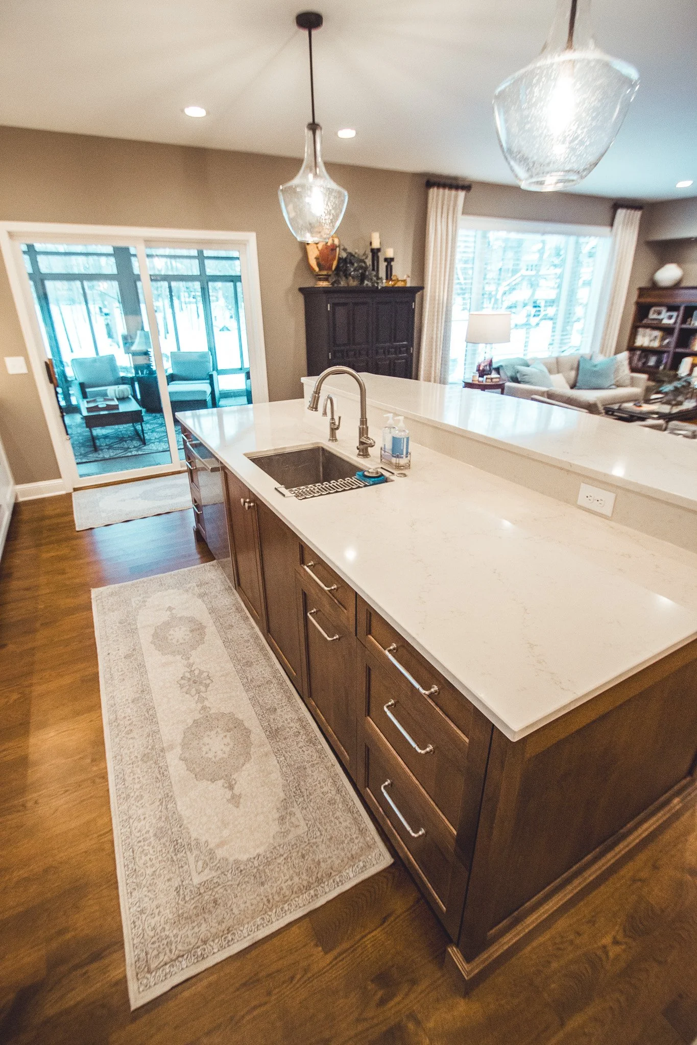Modern kitchen remodel in Chanhassen with wooden cabinets, marble countertop, and pendant lighting. Features a built-in sink, decorative rug on hardwood floor, and view of living area with large window.