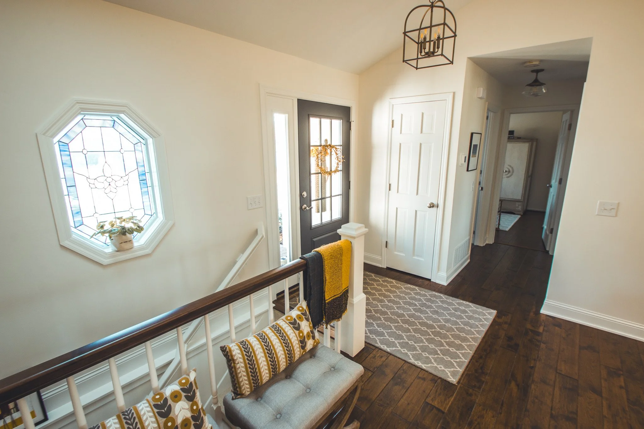 Bright entryway remodel in Minnetonka with a black door, white walls, and hardwood flooring. Features a decorative stained glass window, cushioned bench with patterned pillows, a yellow throw blanket, and a geometric-patterned rug.