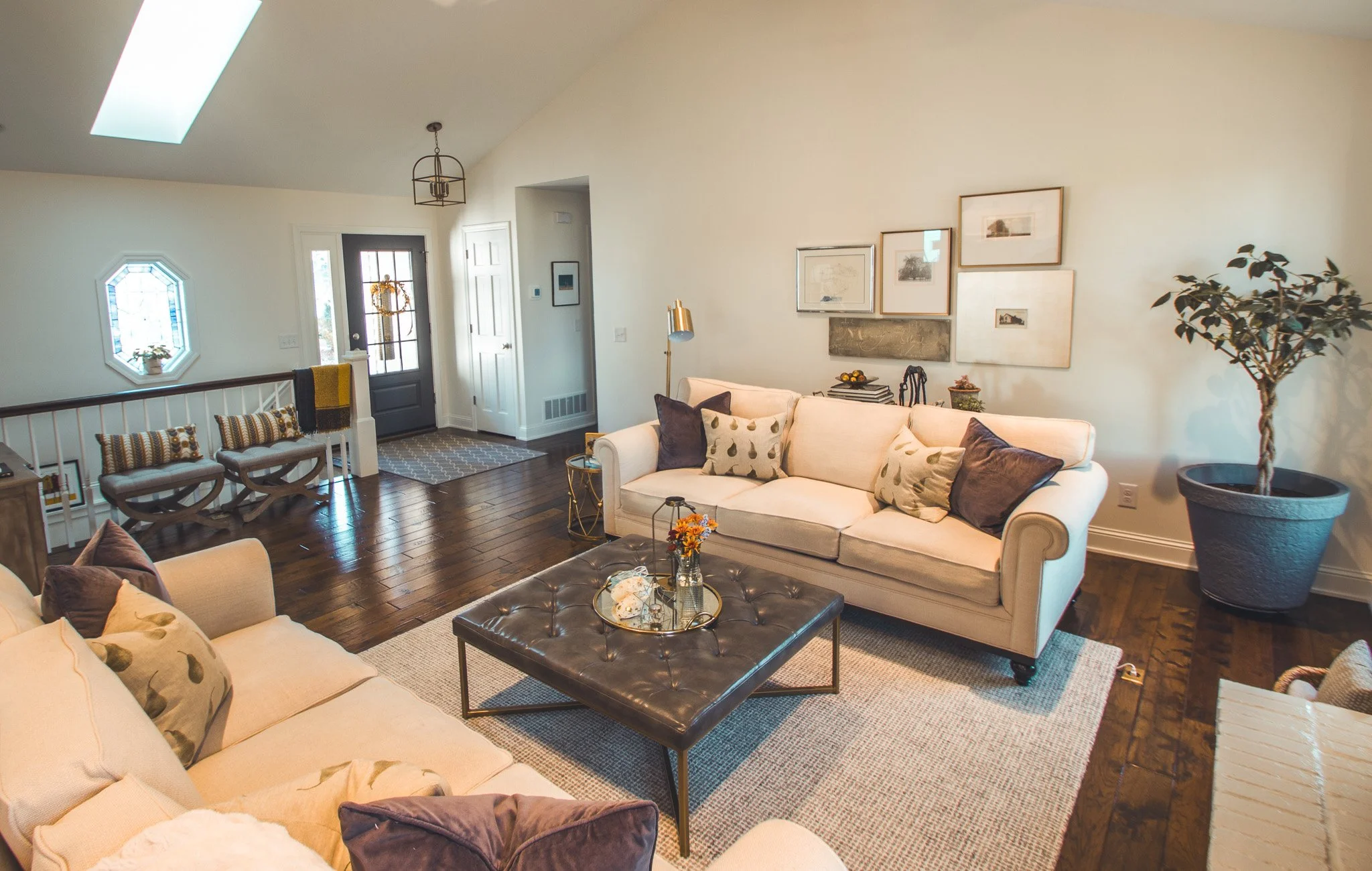 Modern living room remodel in Minnetonka with beige couches, a black tufted coffee table, decorative pillows, framed wall art, and a large potted plant. Wood flooring and natural light from a skylight accentuate the space. 