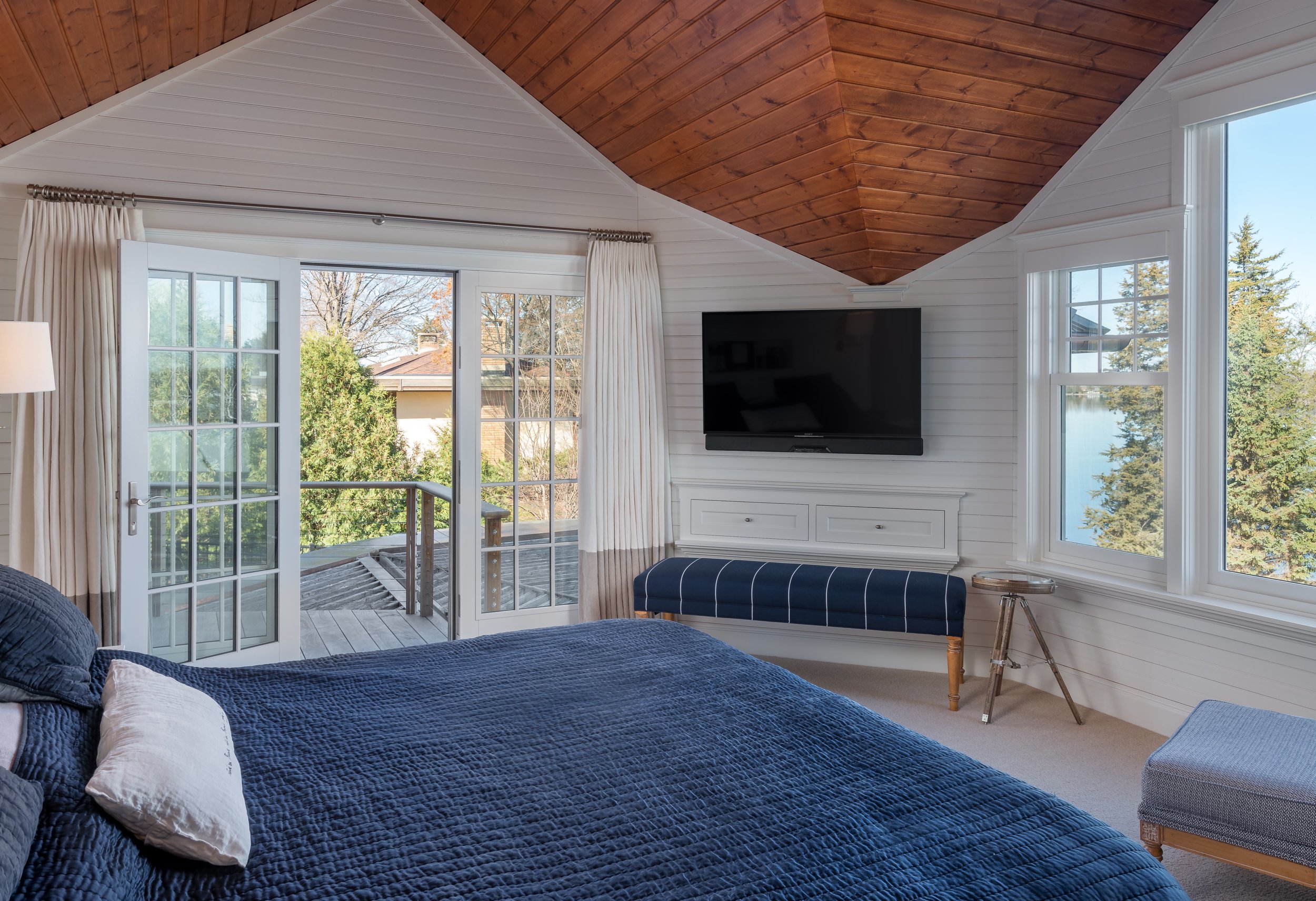 Modern bedroom remodel in Chanhassen with vaulted wood ceiling, large windows, and a sliding glass door leading to a balcony. Features a flat-screen TV, blue bedding, and a bench under the TV.