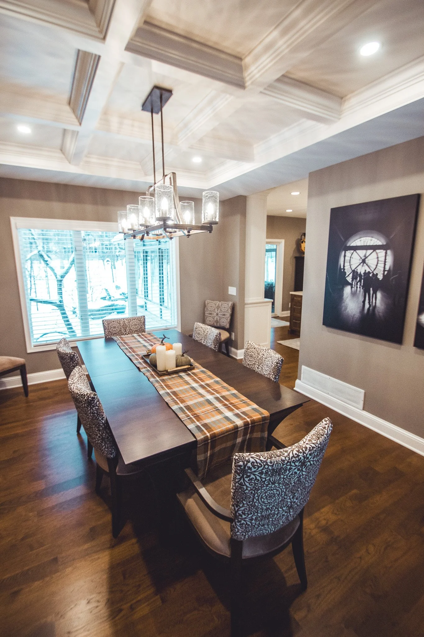 Elegant dining room remodel in Eden Prairie with a dark wood table, cushioned chairs, a plaid table runner, and decorative candles. A chandelier with glass light fixtures hangs from a coffered ceiling, and framed art is on the wall.