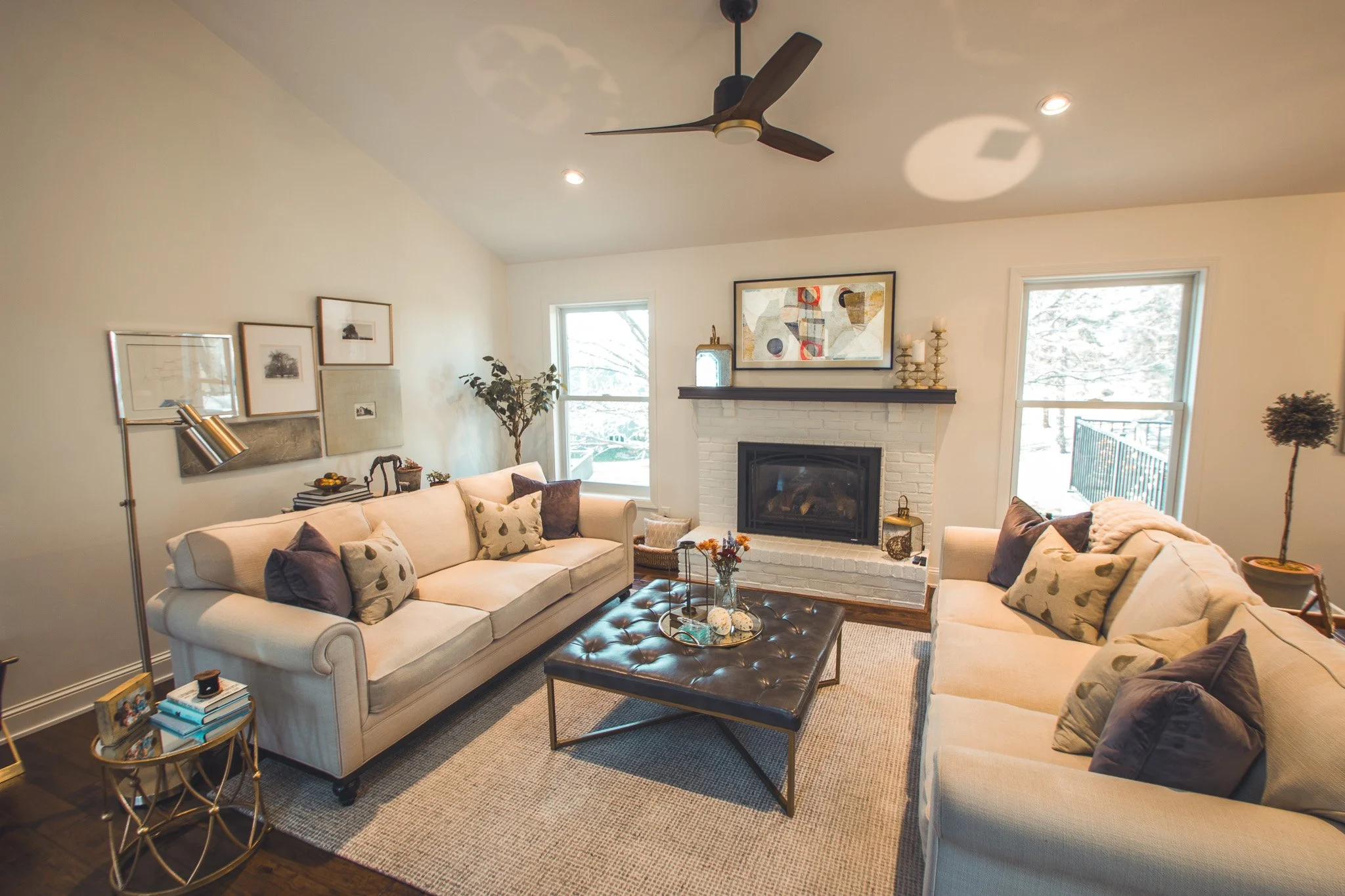 Modern living room remodel in Minnetonka with beige sofas, a black tufted ottoman, white brick fireplace, ceiling fan, and decorative artwork.