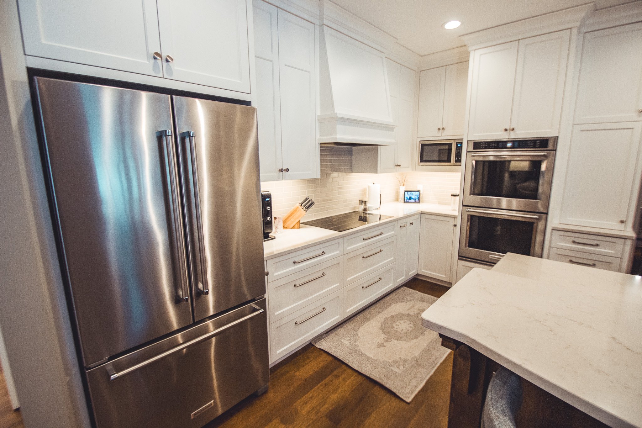 Modern kitchen remodel in Chanhassen with stainless steel refrigerator, white cabinets, double oven, countertop with cutting board and knife set, and a small rug.