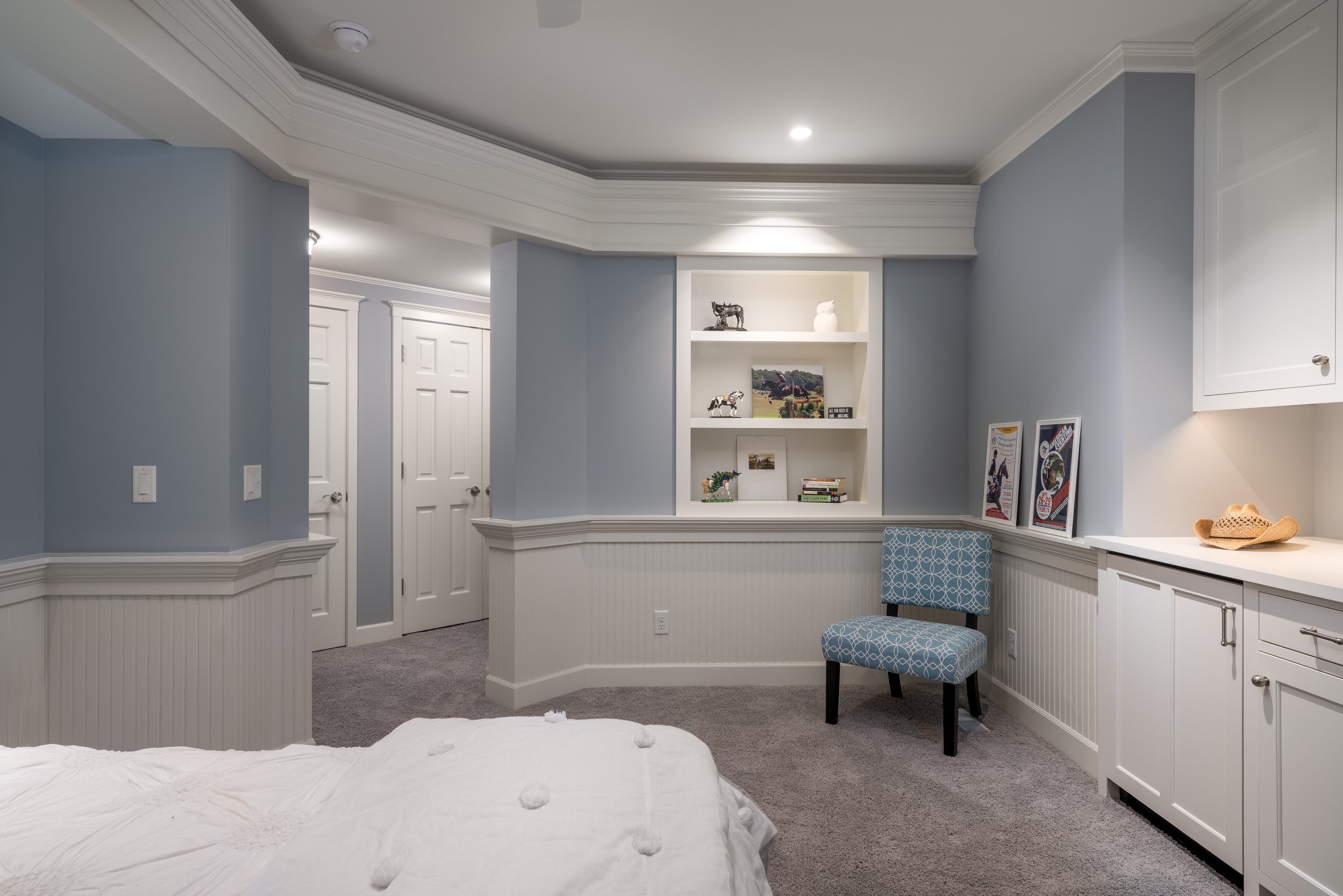 Bedroom remodel in Chanhassen with light blue walls, white trim, built-in shelving, and a blue patterned chair. The shelving displays small decorations and books. A straw hat rests on a countertop beside two framed pictures. 