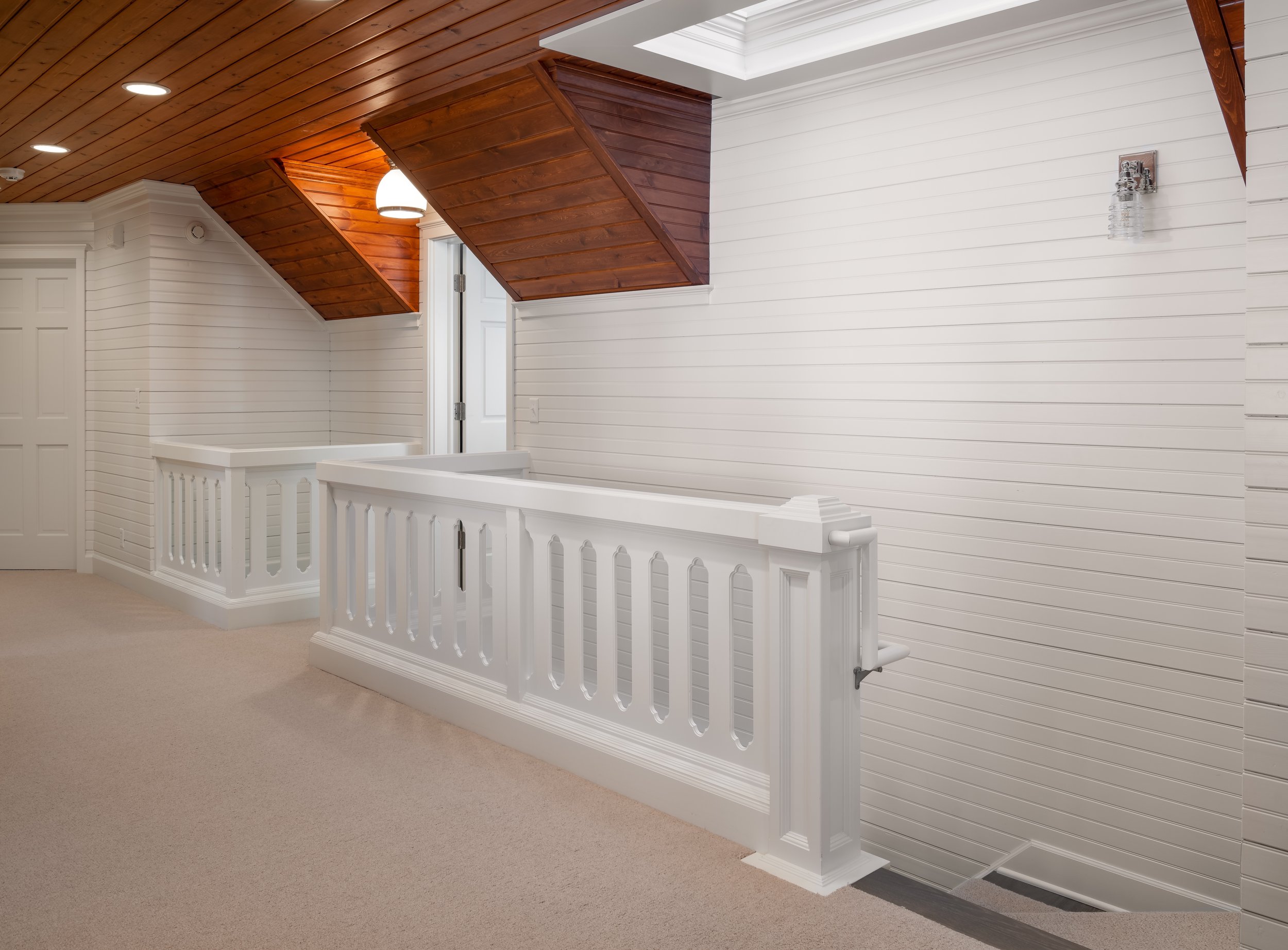 Hallway remodel in Chanhassen with white wooden paneling, a carpeted floor, and a white railing. The ceiling is made of wooden planks with recessed lighting and a skylight. A wall-mounted light fixture is visible.
