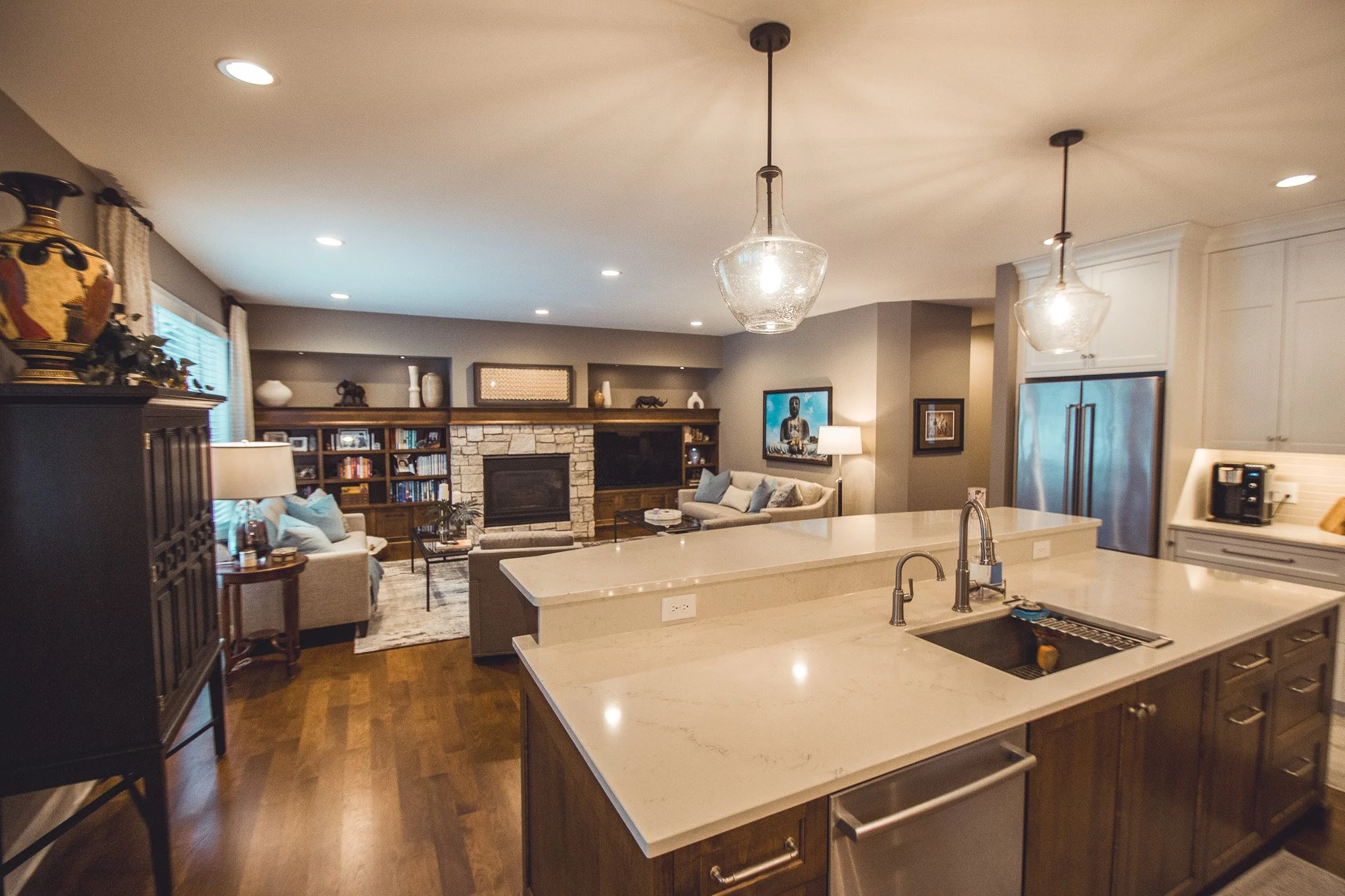 Modern open-concept living room and kitchen in Chanhassen with hardwood floors, stone fireplace, pendant lights, kitchen island, and stainless steel appliances.