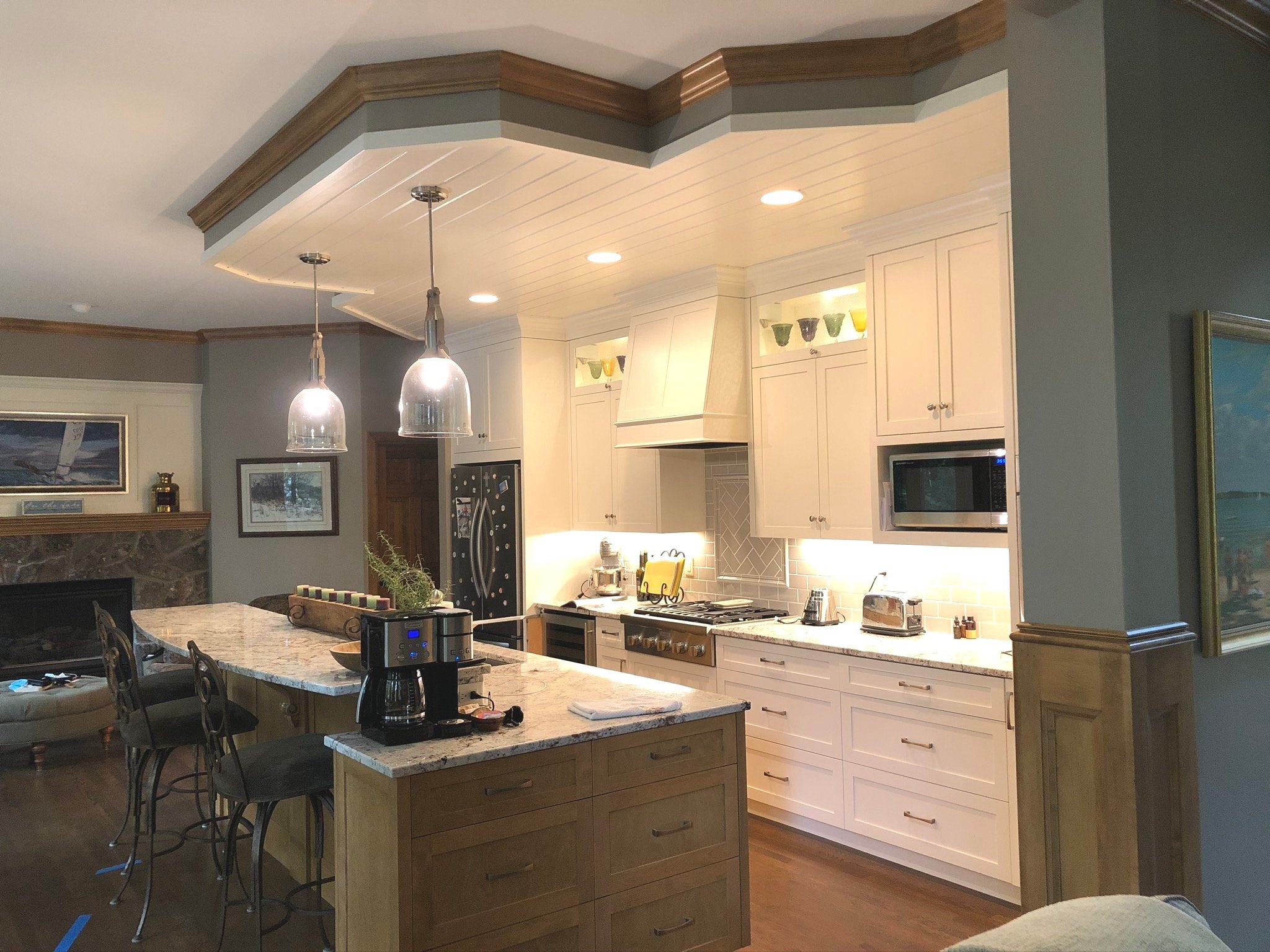 Modern kitchen  in Chanhassen with white cabinets, stainless steel appliances, and a marble island. Two pendant lights hang over the island with barstools. A fireplace and artwork are visible in the background.
