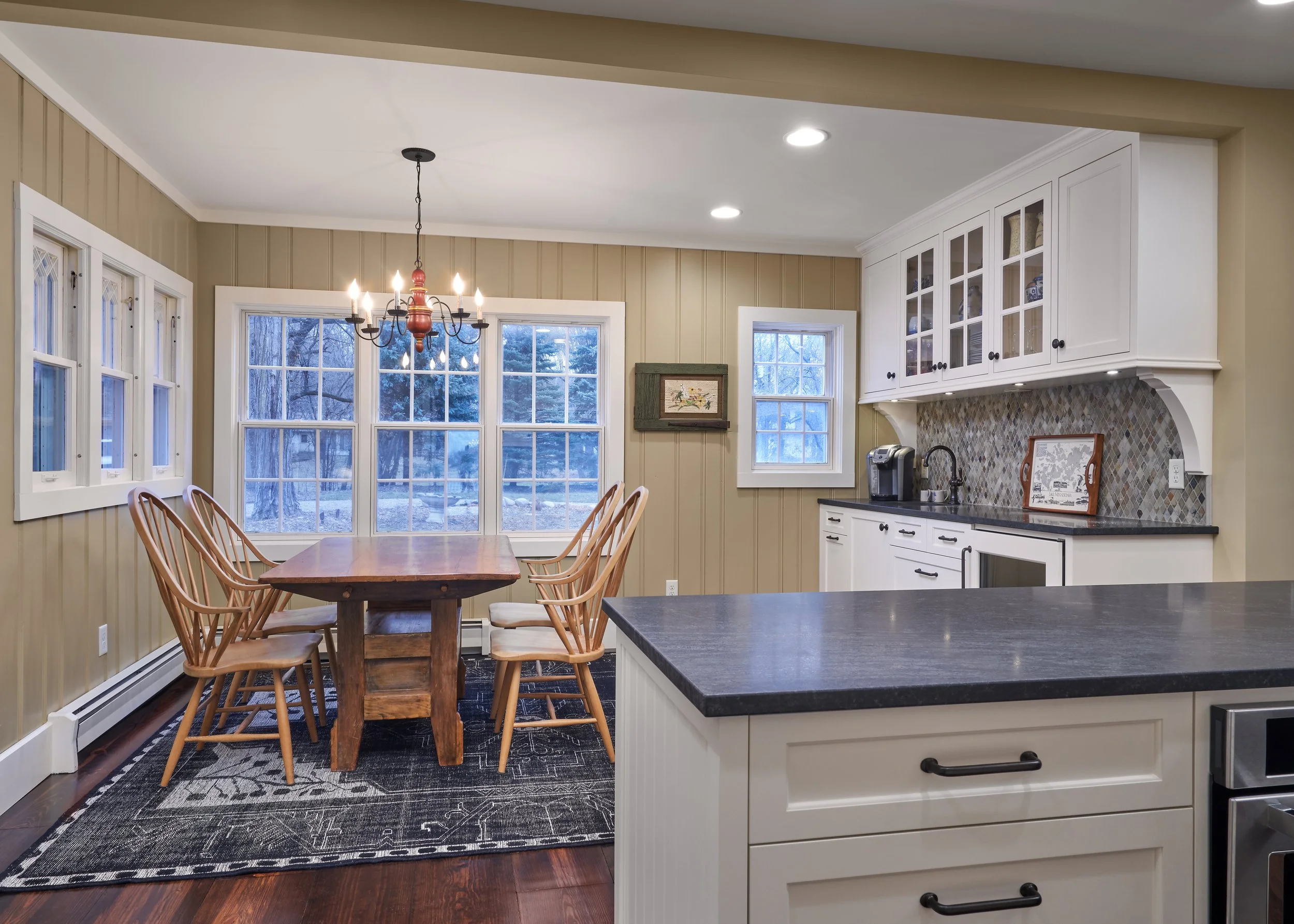 Cozy kitchen and dining area in Chanhassen with wooden table and chairs. The kitchen features white cabinetry, black countertops, patterned backsplash, and a coffee maker.