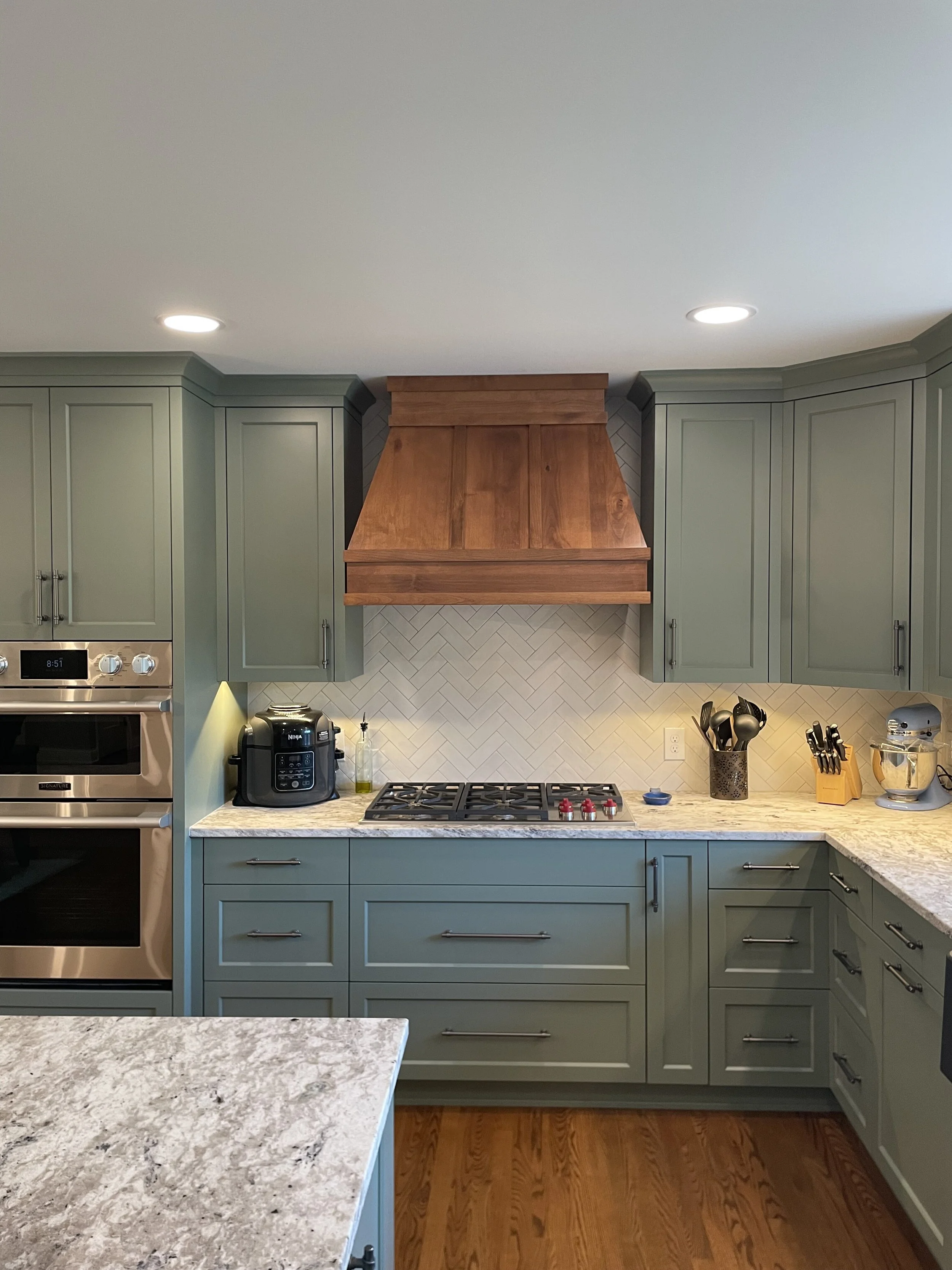 Modern kitchen in Chanhassen with green cabinets, marble countertops, stainless steel double oven, wooden range hood, gas stove, and various kitchen appliances.