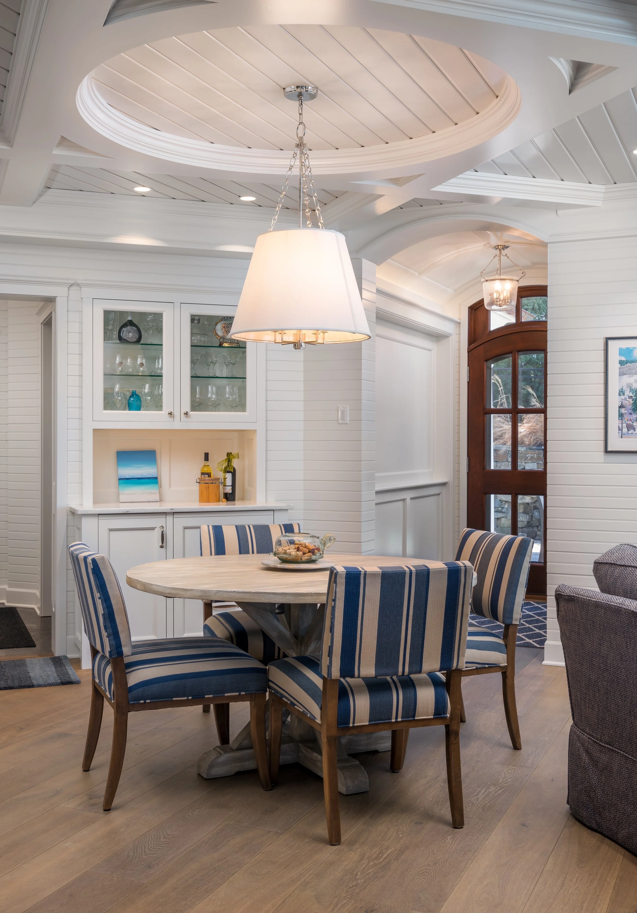Coastal-style dining area on Lake Minnetonka with round wooden table and blue striped chairs, white built-in cabinet with glass doors, and pendant light fixture above. Wooden flooring and arched doorway visible.