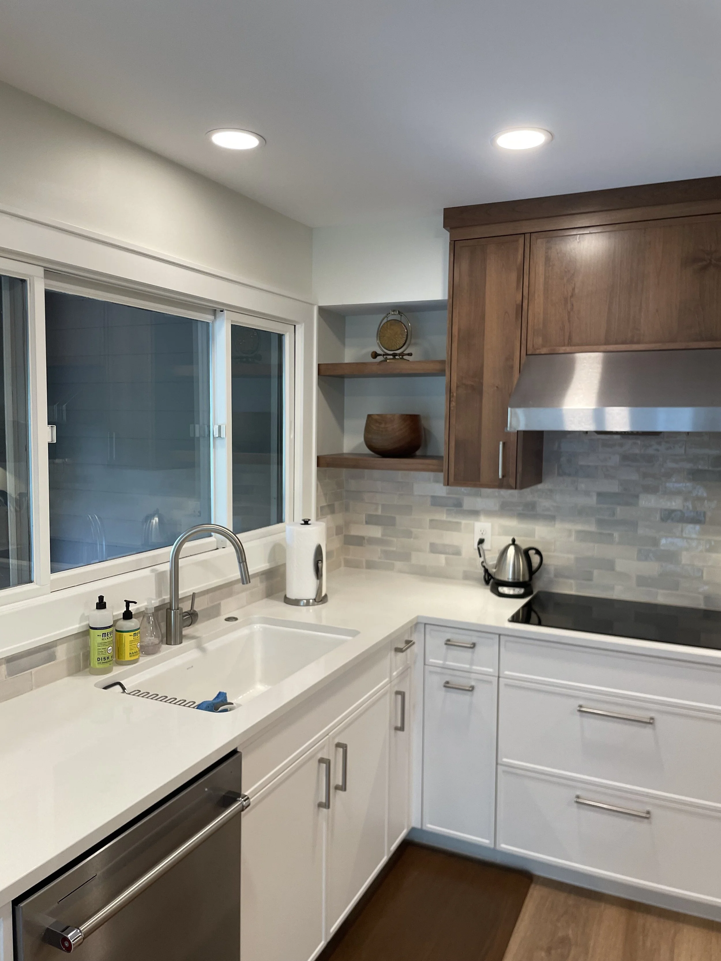 Modern kitchen  in Chanhassen with white cabinetry, stainless steel dishwasher, and electric stove. Features a stainless steel faucet, white sink, paper towel holder, and cleaning supplies on the counter. Wooden shelves hold decorative items. 