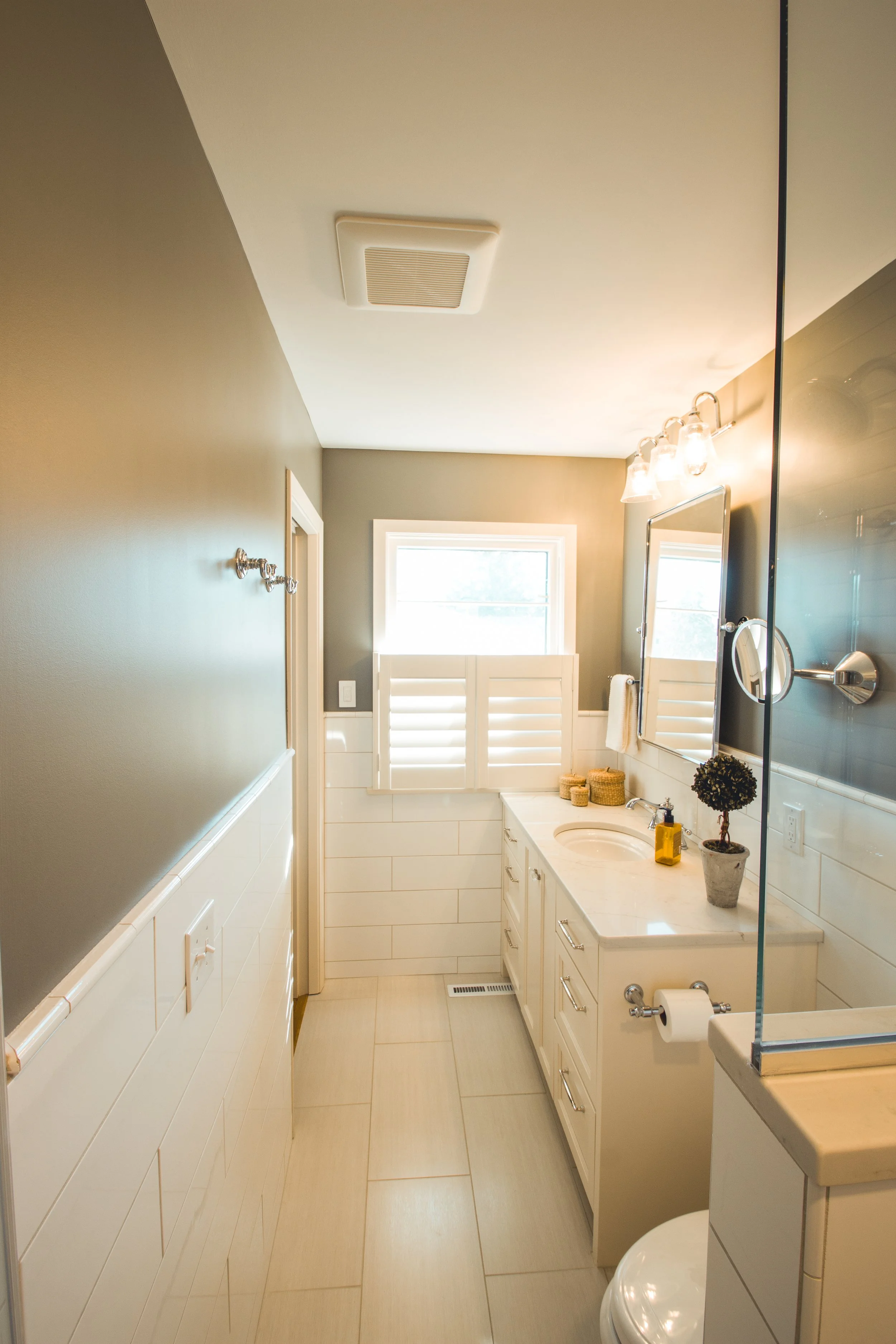 Small modern bathroom remodel in Chanhassen with a white vanity, beige tiles, a window with shutters, and a mirror above the sink.