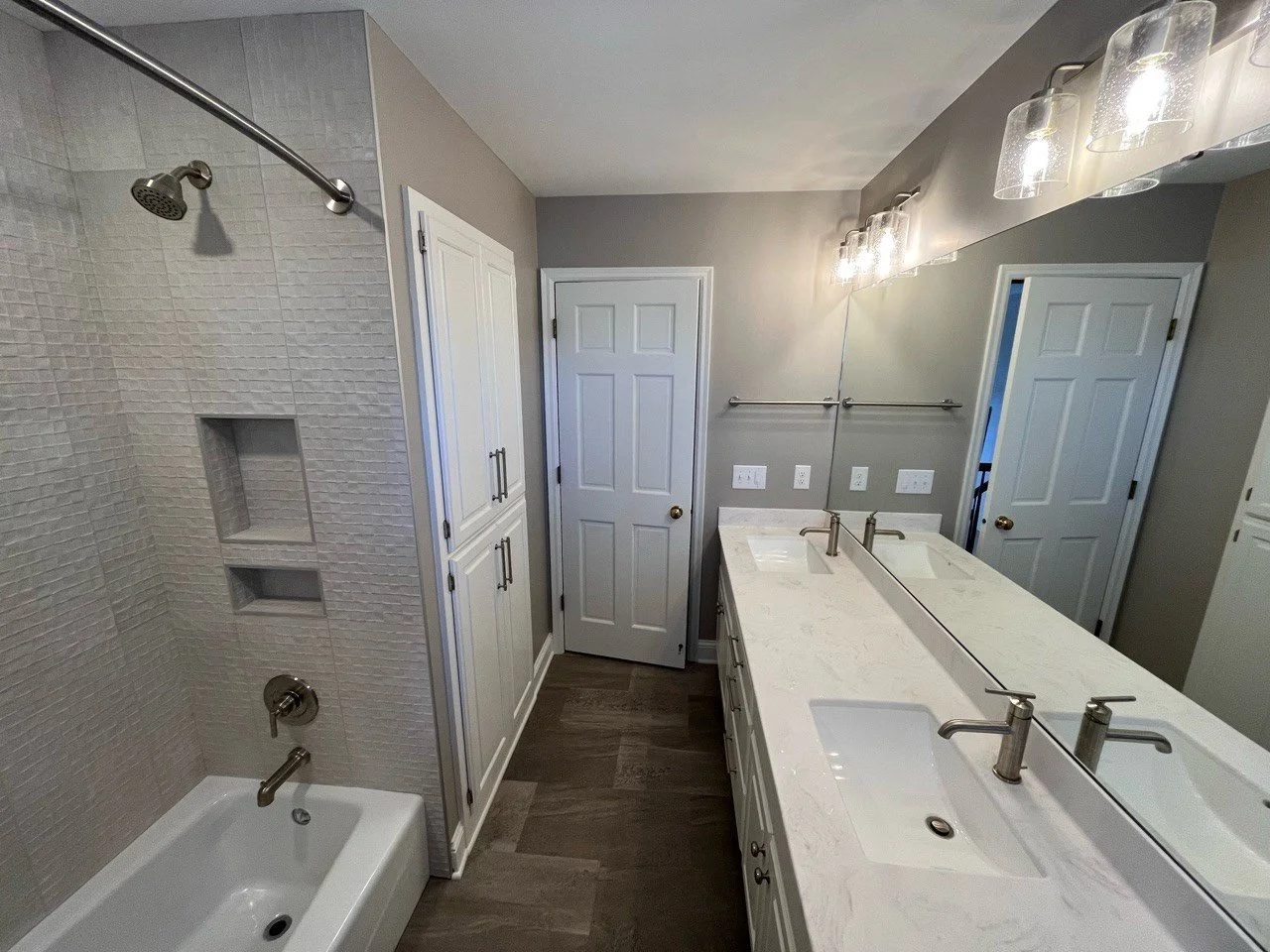 Modern bathroom remodel in Eden Prairie with white double sink vanity and large mirror, gray tiled shower with built-in shelves and chrome fixtures, light gray walls, and overhead lighting.