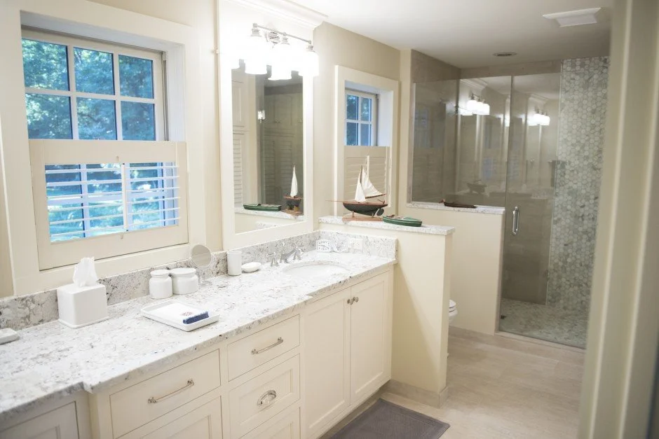 Modern bathroom remodel in Chanhassen with white marble countertop, dual sinks, and glass shower. Features include storage cabinets, sailboat decoration, and a window with shutters.