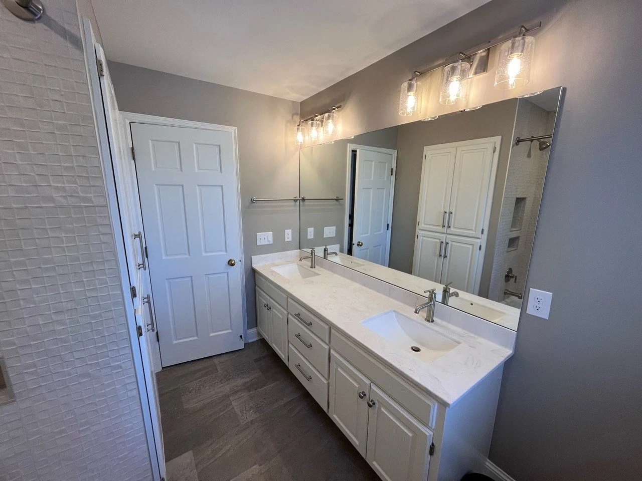 Modern bathroom remodel in Chanhassen with white vanity, double sinks, large mirror, gray walls, overhead lighting, and a closed door.