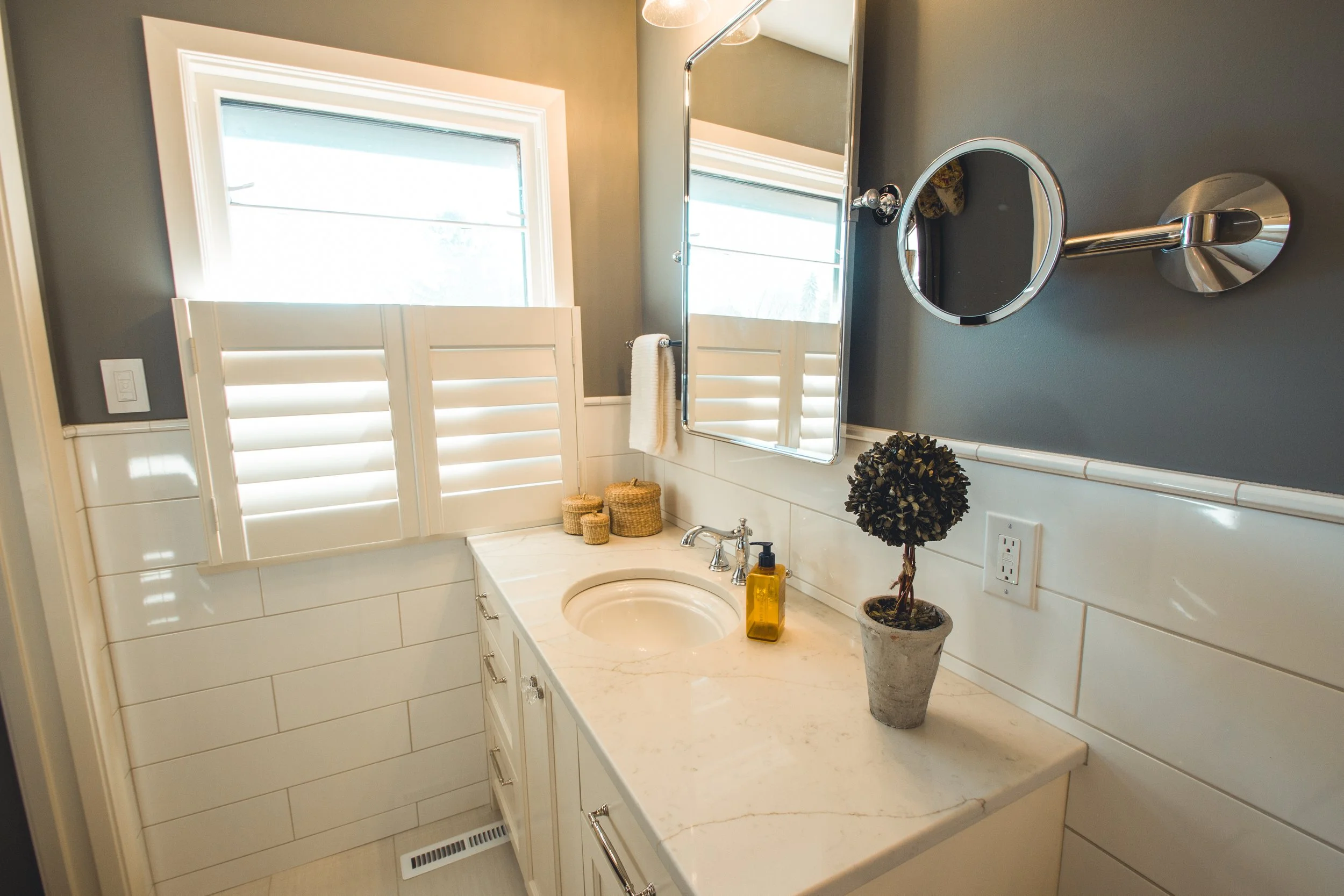 A bathroom remodel in Chanhassen with a white countertop, sink, and faucet. There's a potted plant, soap dispenser, and wicker baskets on the countertop. A mirror, circular vanity mirror, and towel hang on the wall. Shutters cover a window partially.