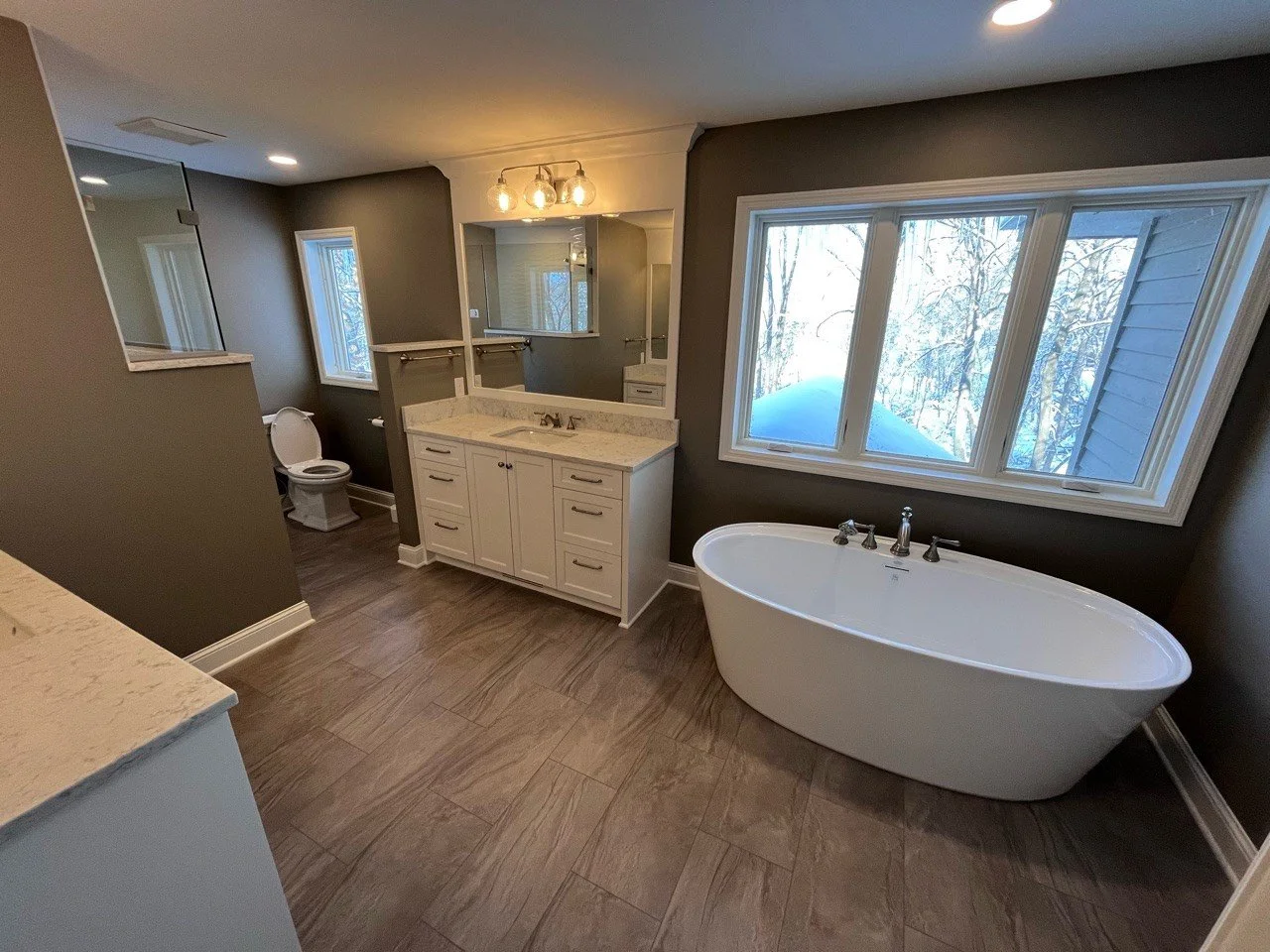 Modern bathroom remodel in Chanhassen with freestanding bathtub, large windows, double-sink vanity, and toilet, featuring wood-look tile flooring and neutral wall colors.