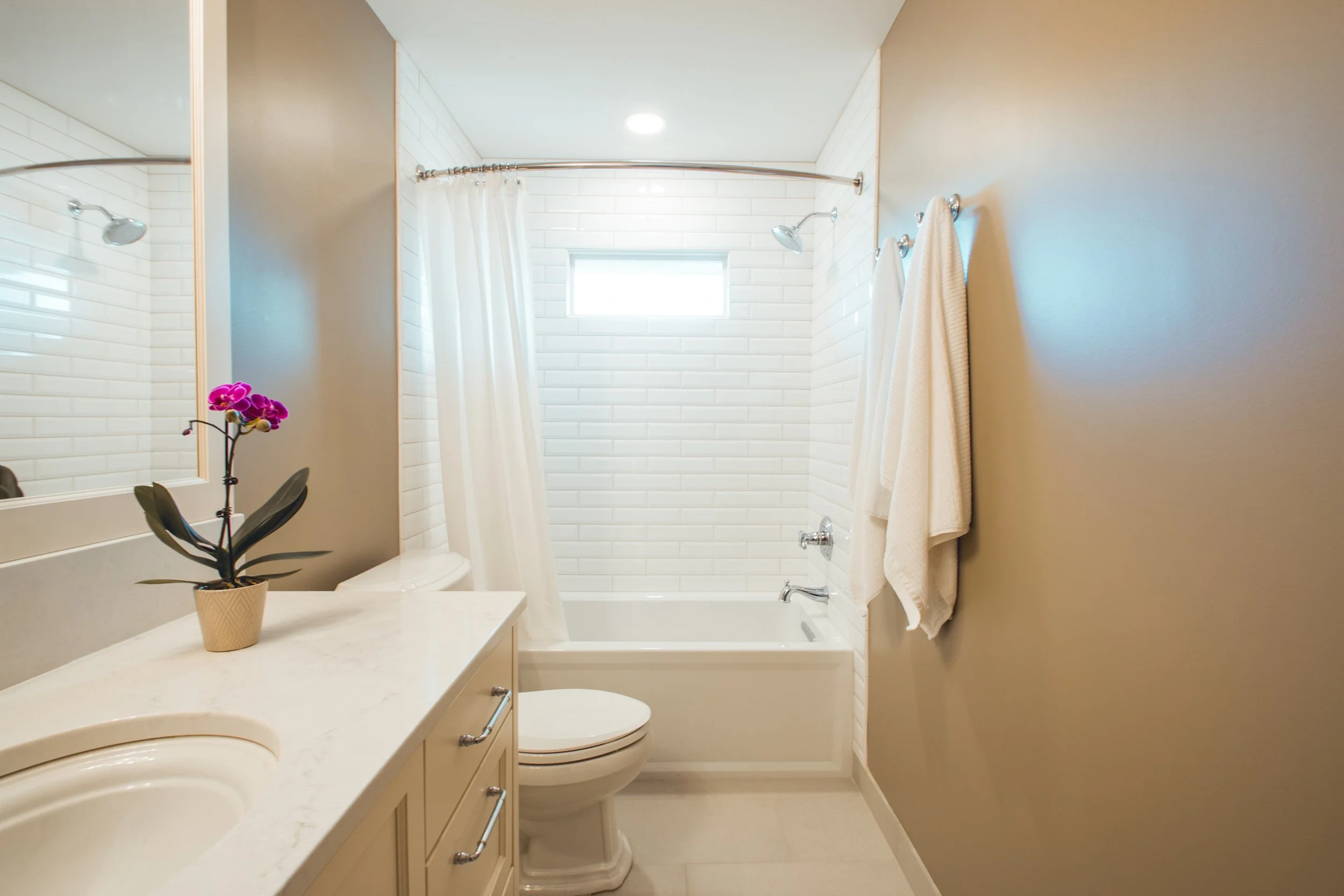 Modern bathroom remodel in Chanhassen with a white bathtub, shower curtain, tiled walls, sink with countertop, orchid in a pot, and towels hanging on the wall.