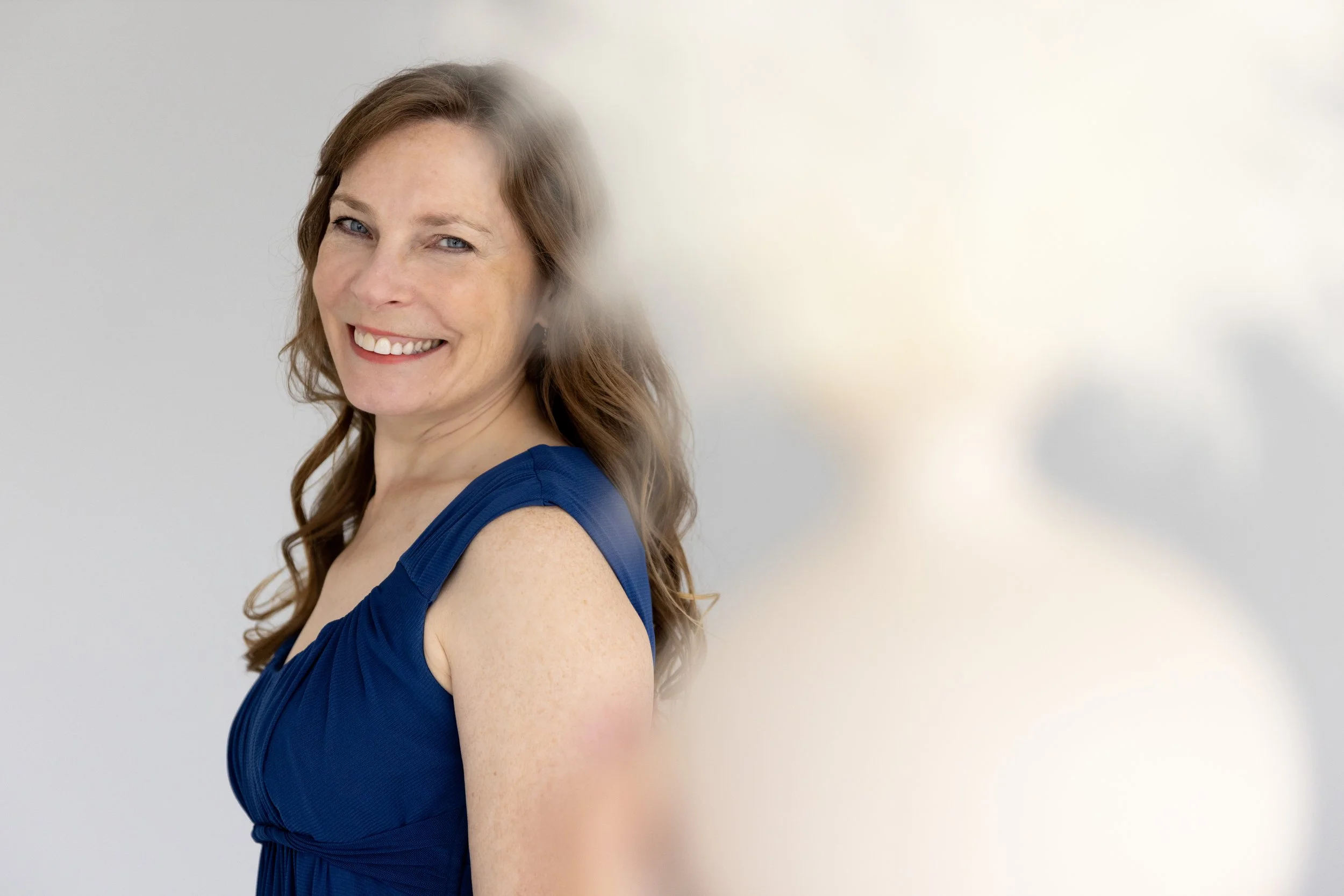 A woman with long brown hair, wearing a blue sleeveless dress, smiling, with an out-of-focus bright background.