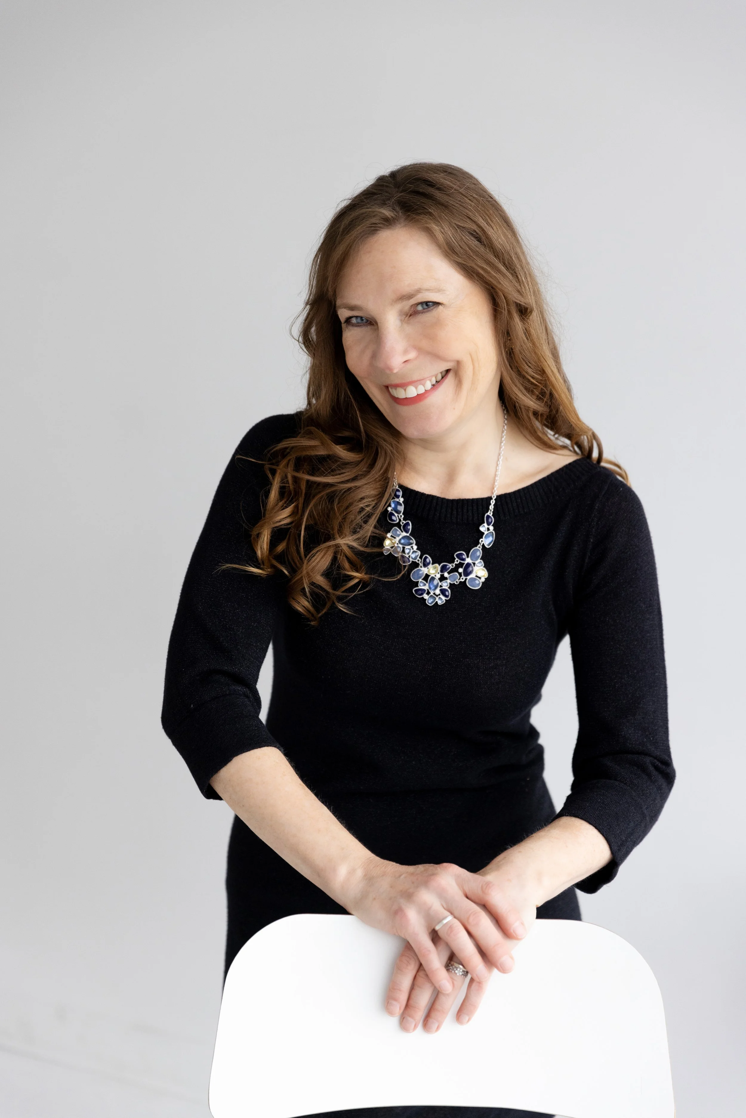 A woman with long brown hair smiling, wearing a black top and a statement necklace, leaning on a white chair against a plain gray background.