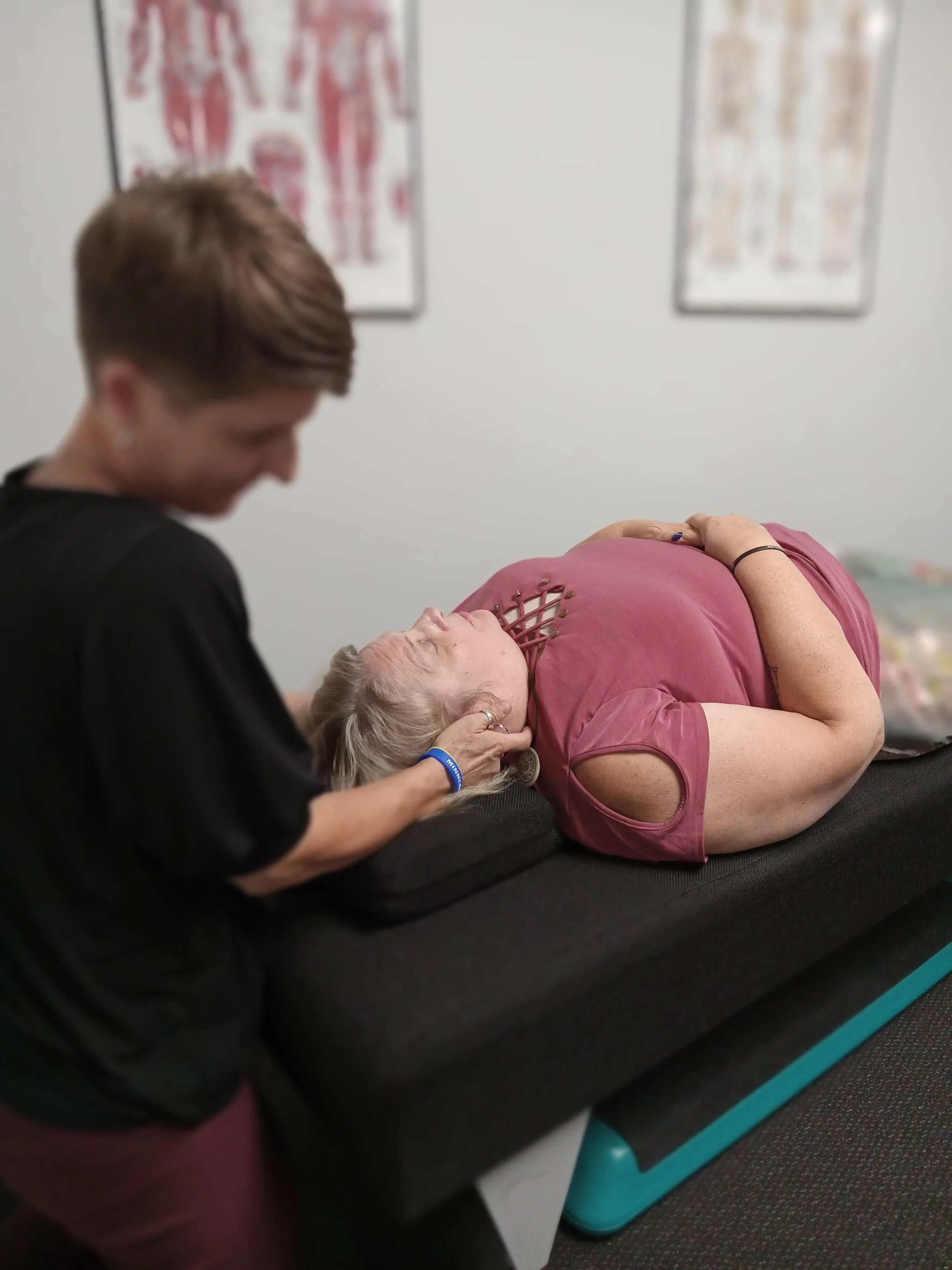 A female chiropractor performing a neck stretches on a female patient lying on an adjusting table in a clinical setting with anatomical posters on the wall.