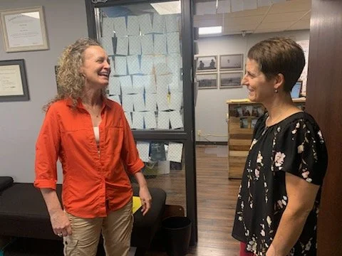 Two women laughing, smiling and talking in an office. One woman is wearing an orange shirt, and the other is wearing a black floral top.