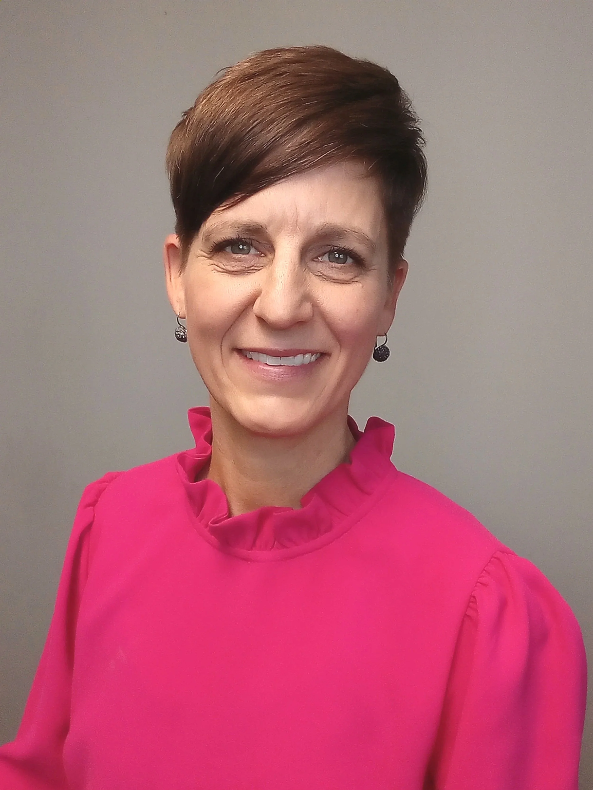 Woman with short brown hair and green eyes wearing a bright pink blouse with a ruffled color and black earrings