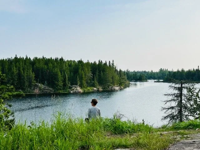 A woman sitting on grass near a lake in northern MN surrounded by pine trees under a clear sky.