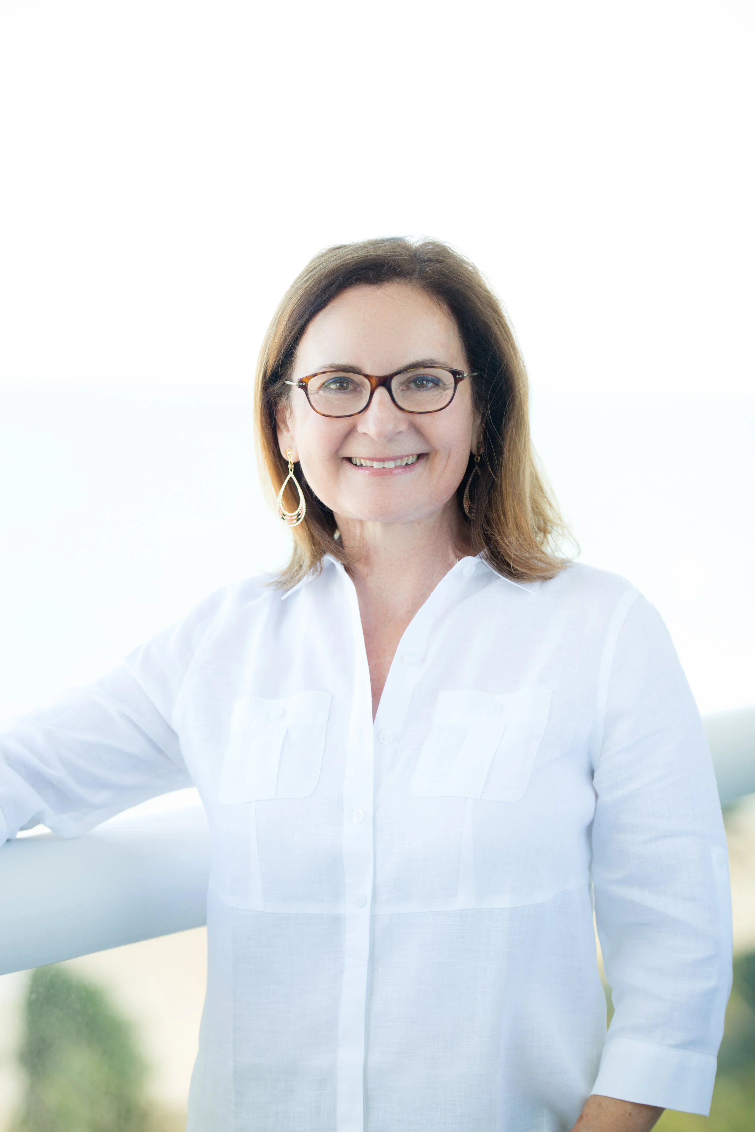 A woman with shoulder-length brown hair, glasses, and earrings, smiling outdoors in a white blouse against a blurred background of greenery.