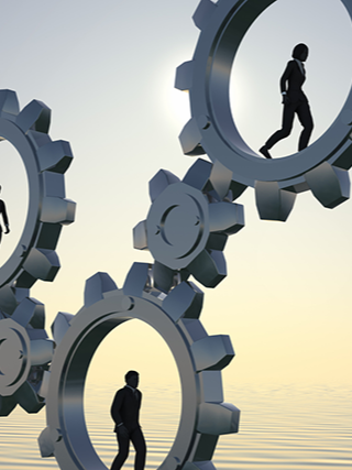 Silhouetted figures walking inside large interconnected gears against a sunrise or sunset background.