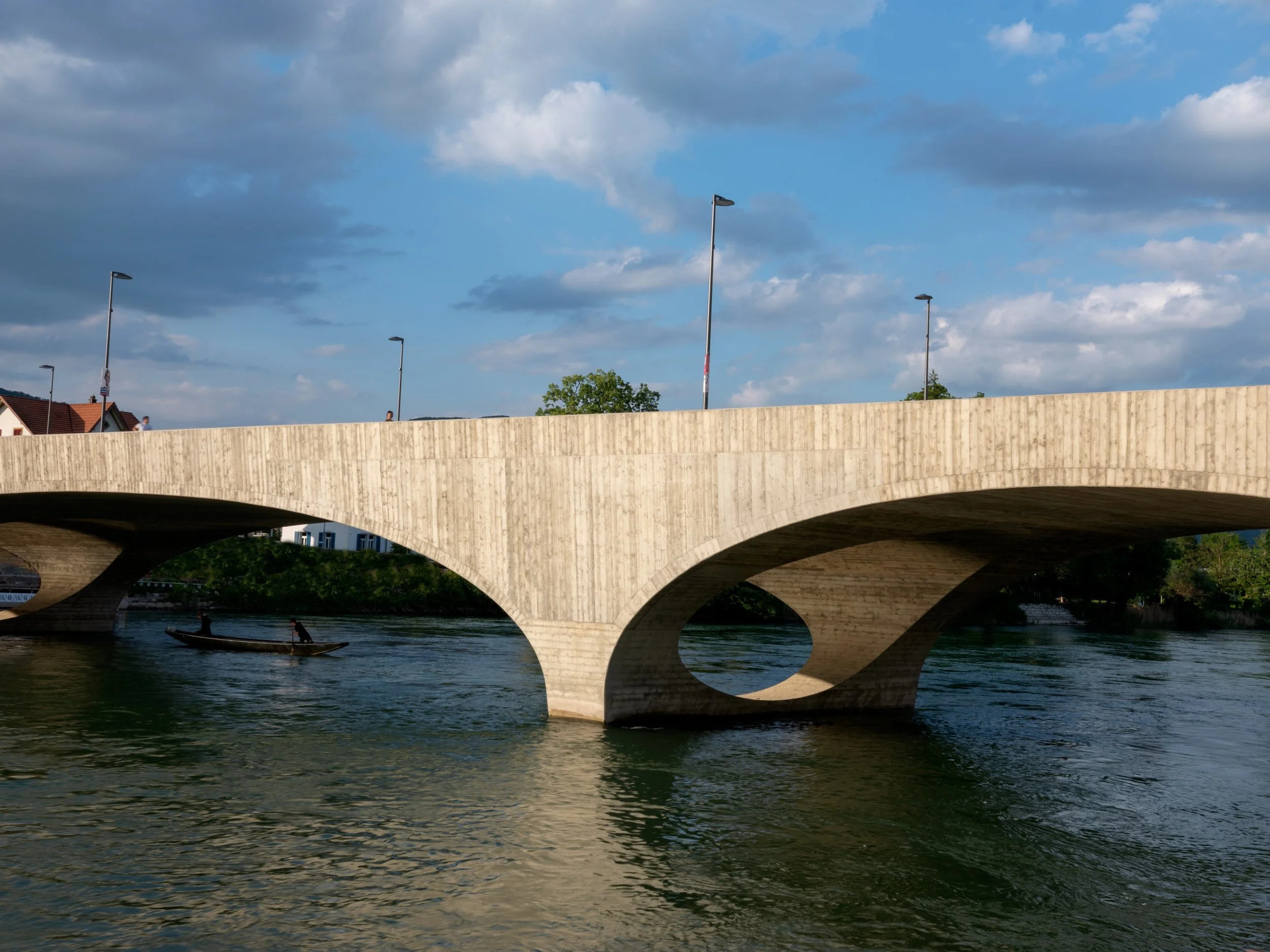 A modern concrete bridge over a river with a person riding a boat underneath. The sky has some clouds.