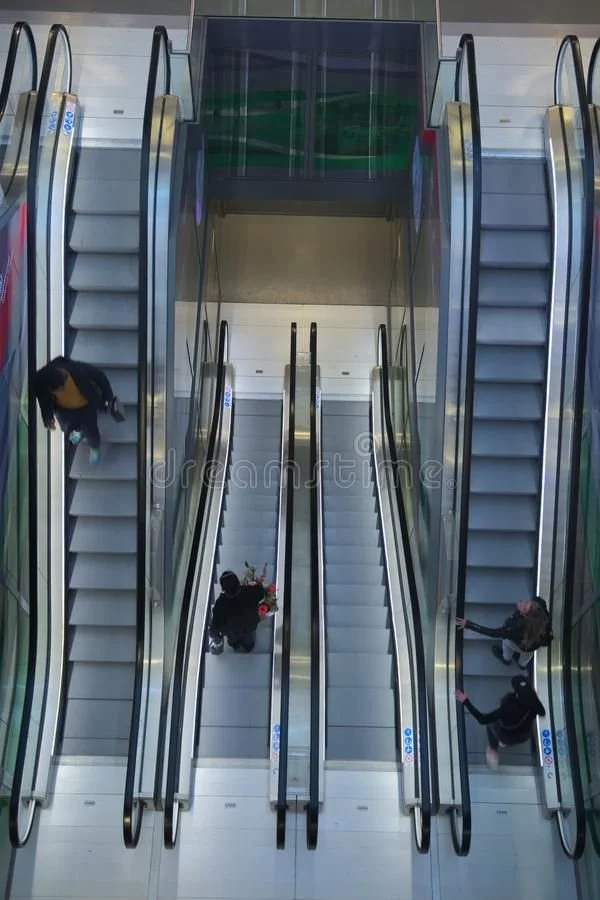 View of multiple escalators inside a shopping mall from an overhead perspective, with several people riding and walking around.