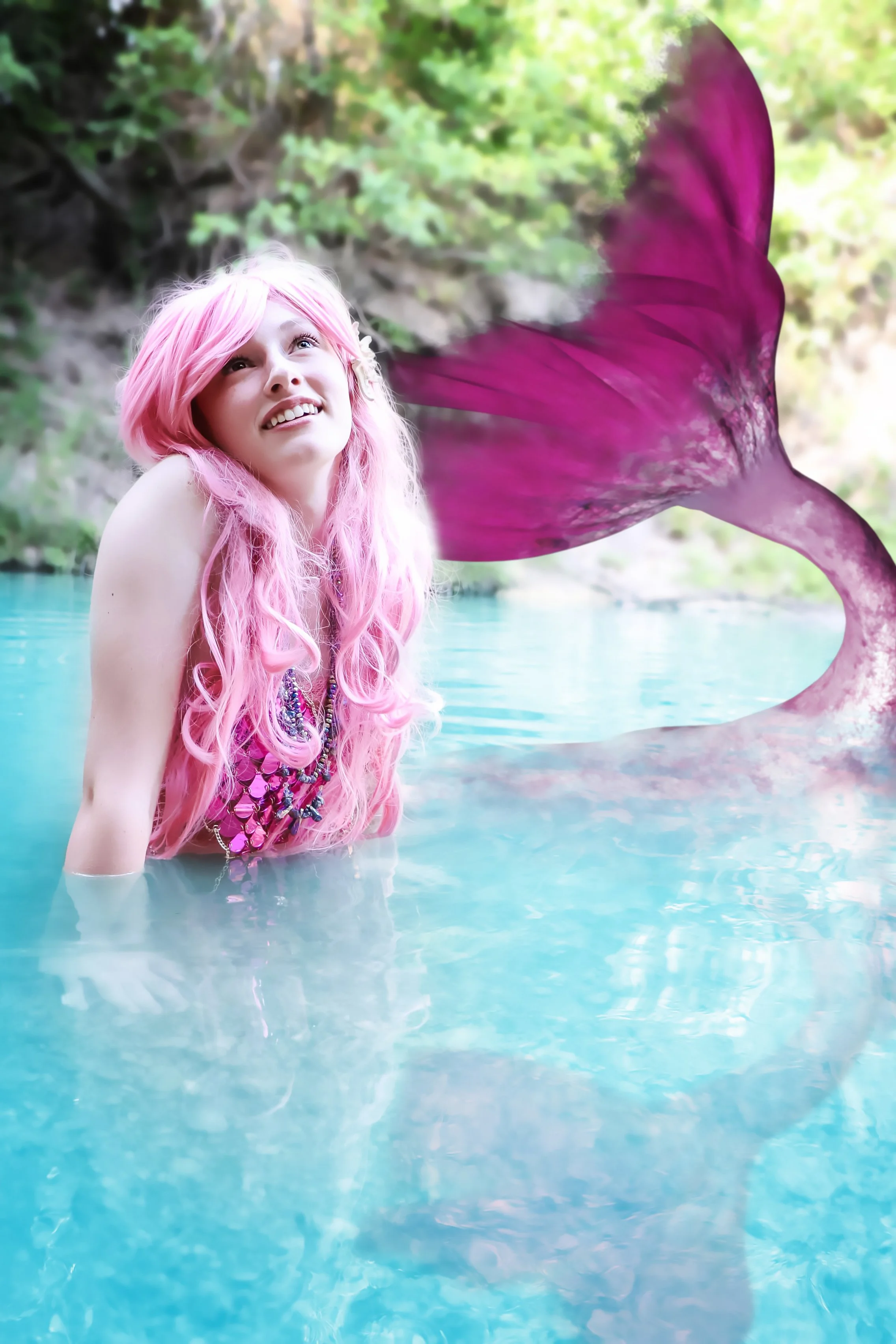 A woman with long pink hair in a water body with a purple mermaid tail, surrounded by greenery in Salado, Texas.