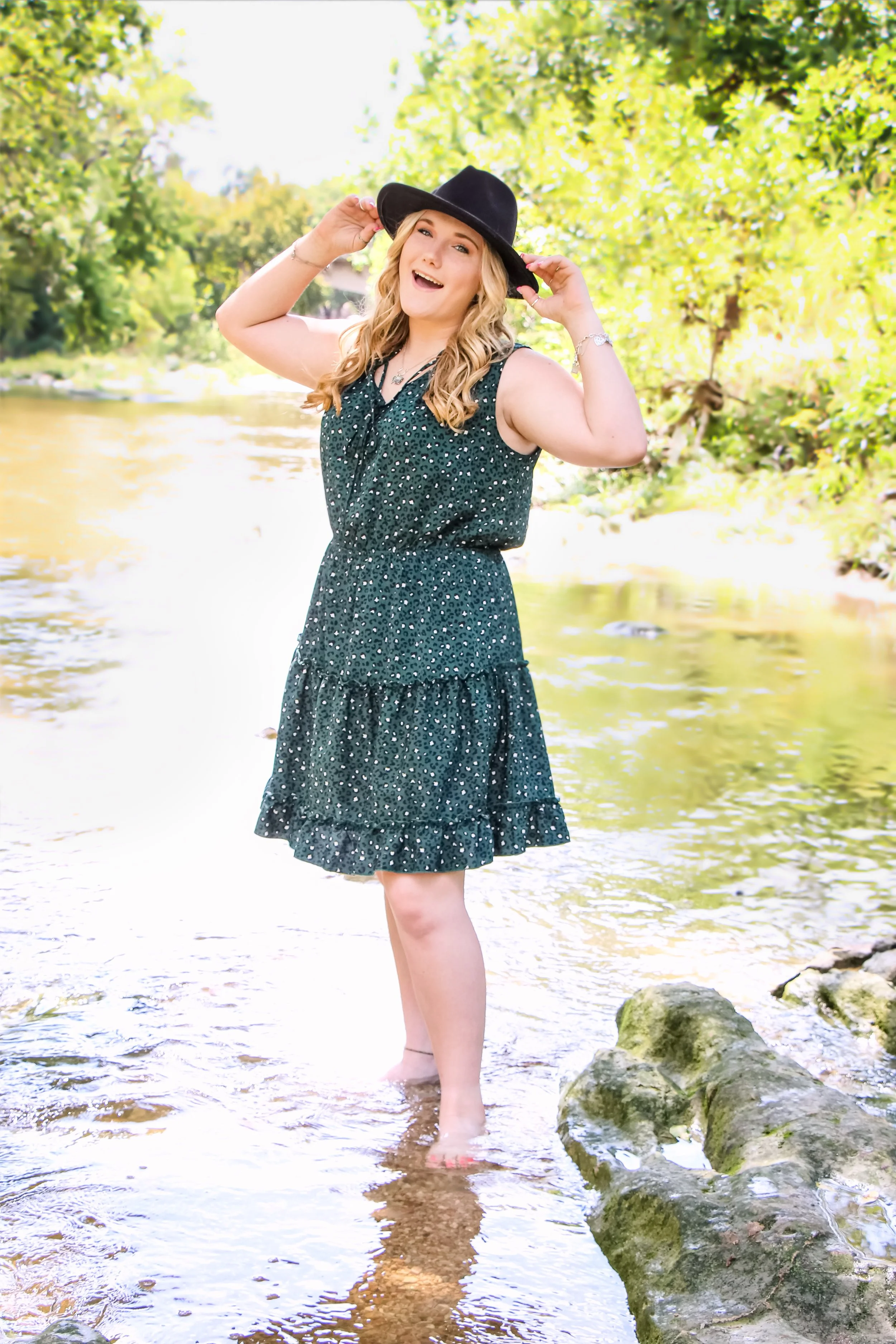 A young woman with blonde hair wearing a black hat and a sleeveless dark green dress, standing in a shallow river during daytime surrounded by trees with yellow and green leaves, smiling and holding her hat.