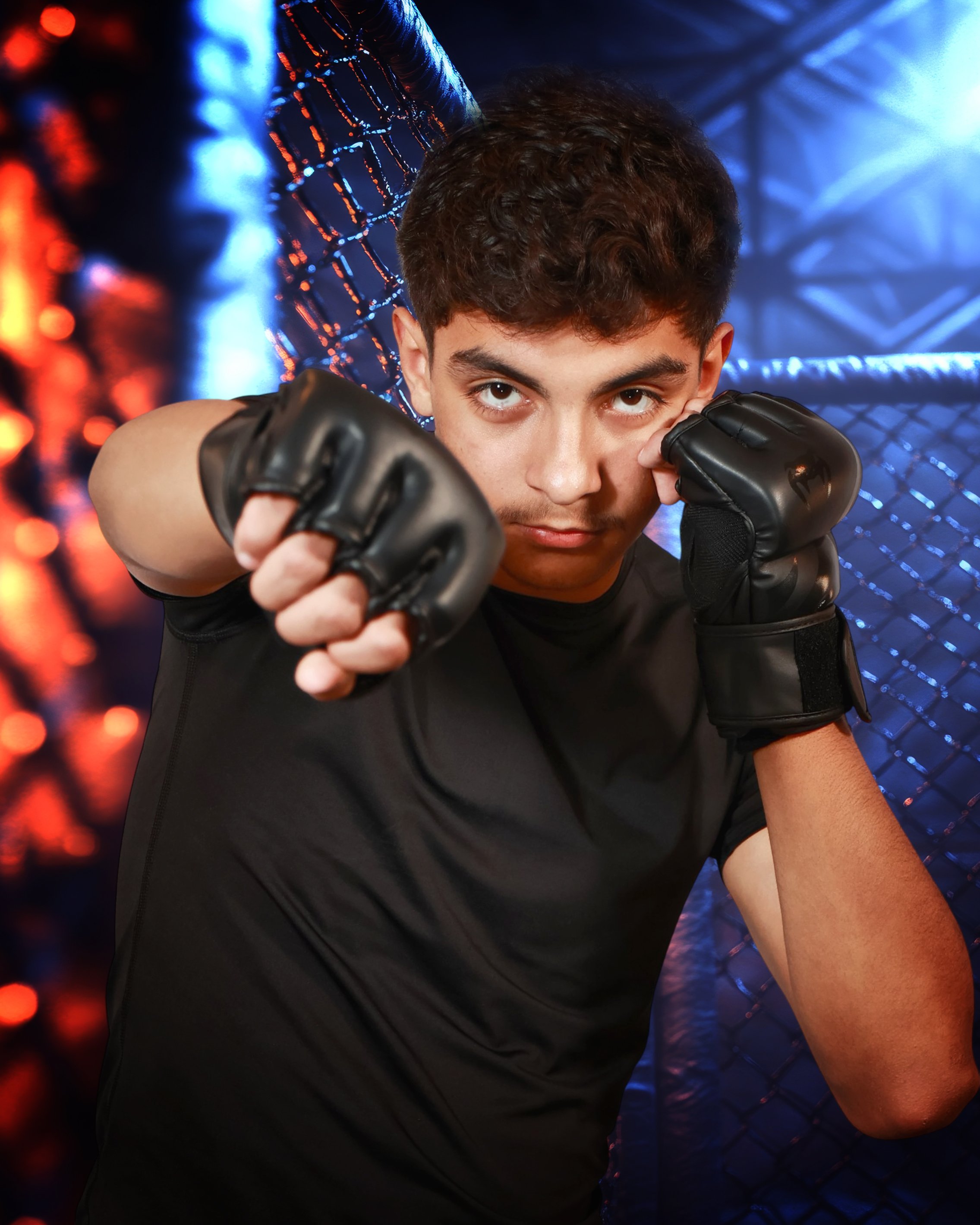Young male mixed martial artist in black shirt and gloves in fighting stance inside an octagon cage with blue and red lighting.
