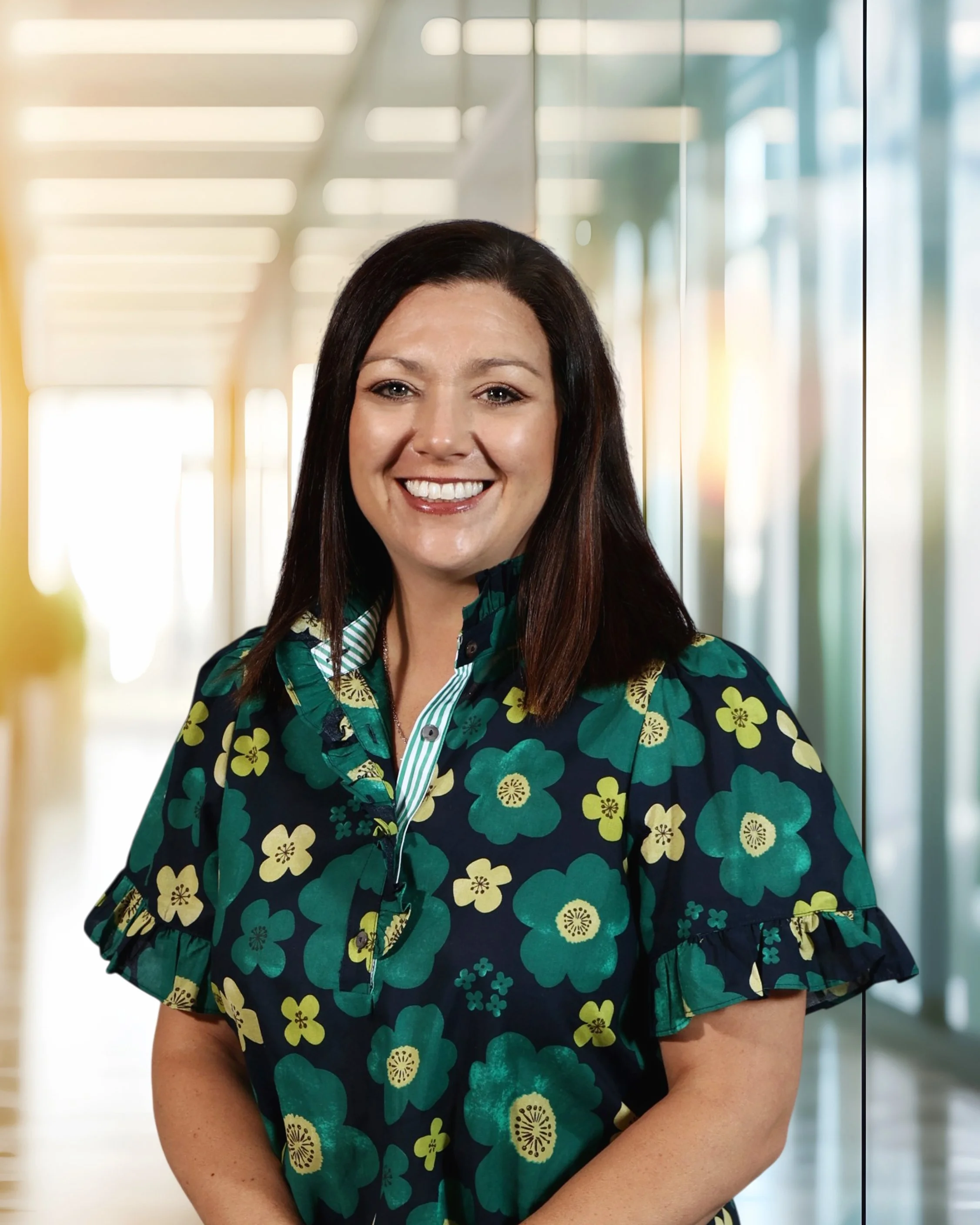 A woman with shoulder-length dark hair, wearing a dark blue floral print blouse, smiling in a modern office hallway with glass walls and bright lighting.