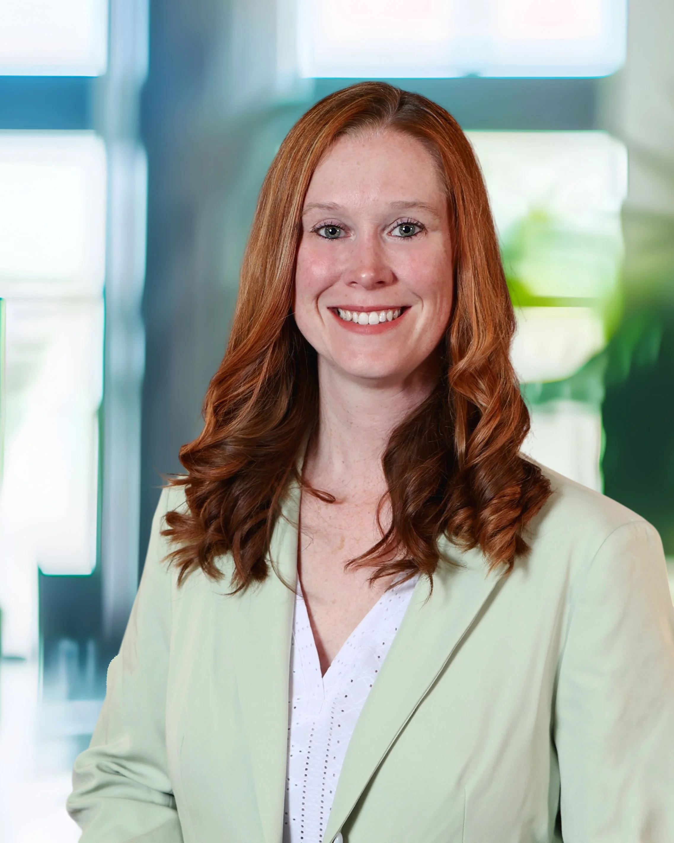 A woman with long red hair smiling, wearing a light-colored blazer and white blouse, in an office setting with large windows and blurred background.