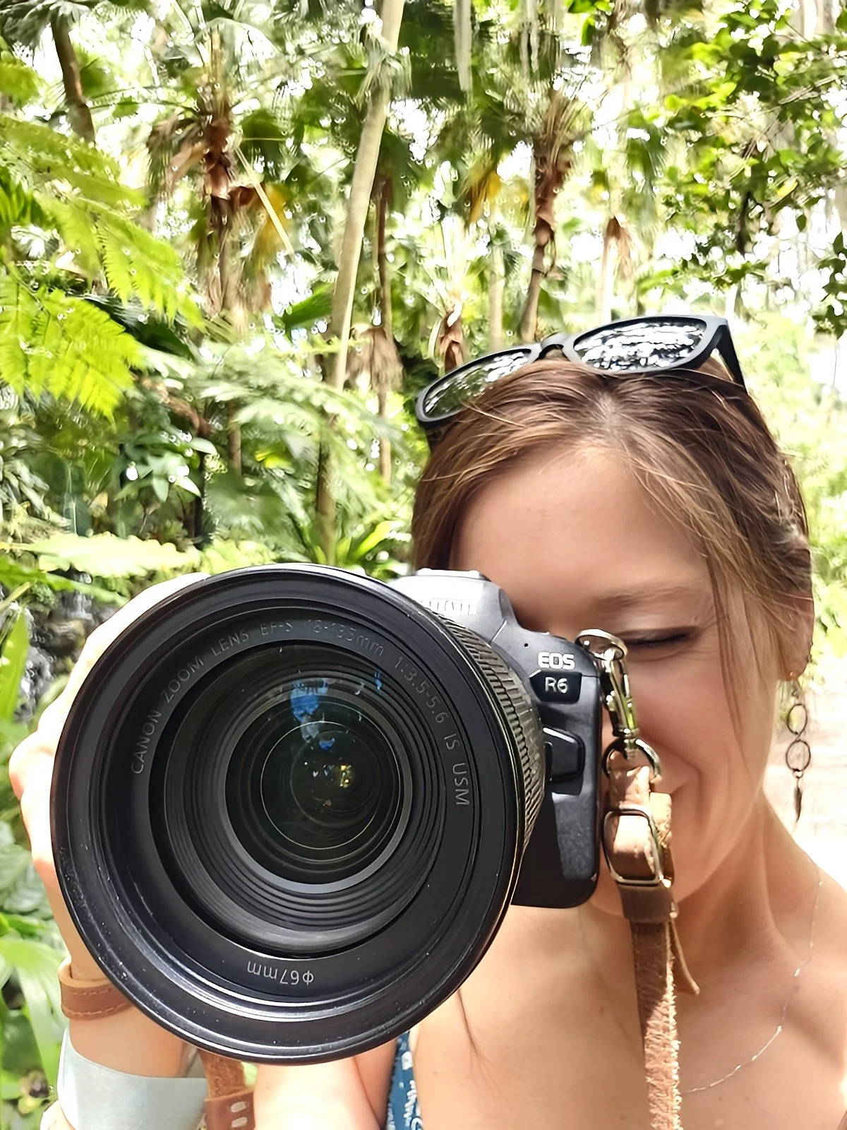 Megan Gibson taking a photo with a Canon EOS R6 camera in a lush green tropical environment, with sunglasses on her head and her eye closed while aiming the camera.