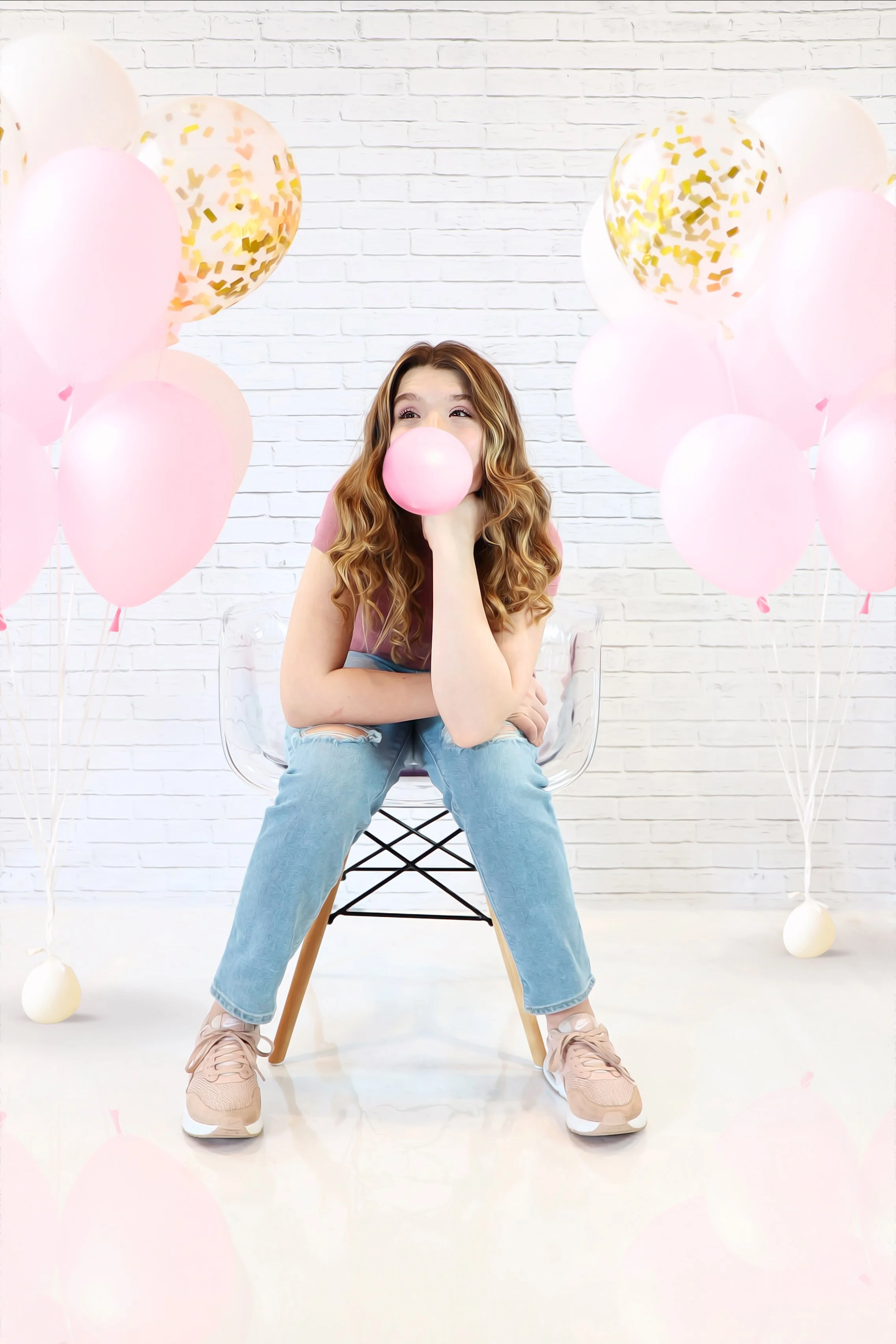A quirky young woman sitting on a transparent chair, blowing a pink bubble gum bubble, surrounded by pink and white balloons with some gold confetti, against a white brick wall.