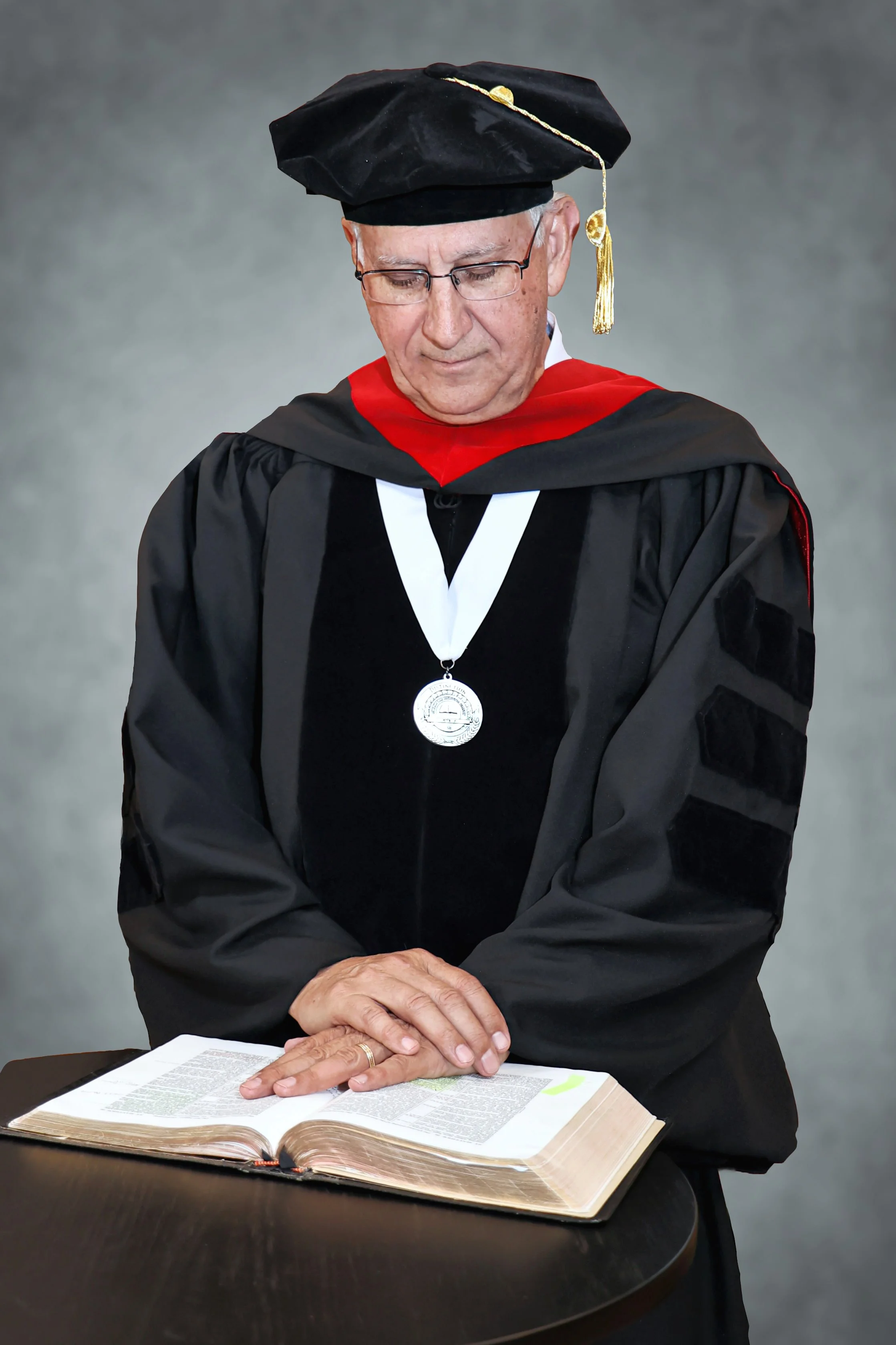 An elderly man dressed in graduation regalia, including a black cap with gold tassel, black gown with red and black accents, medallion, and glasses, standing over an open Bible on a table with hands clasped.