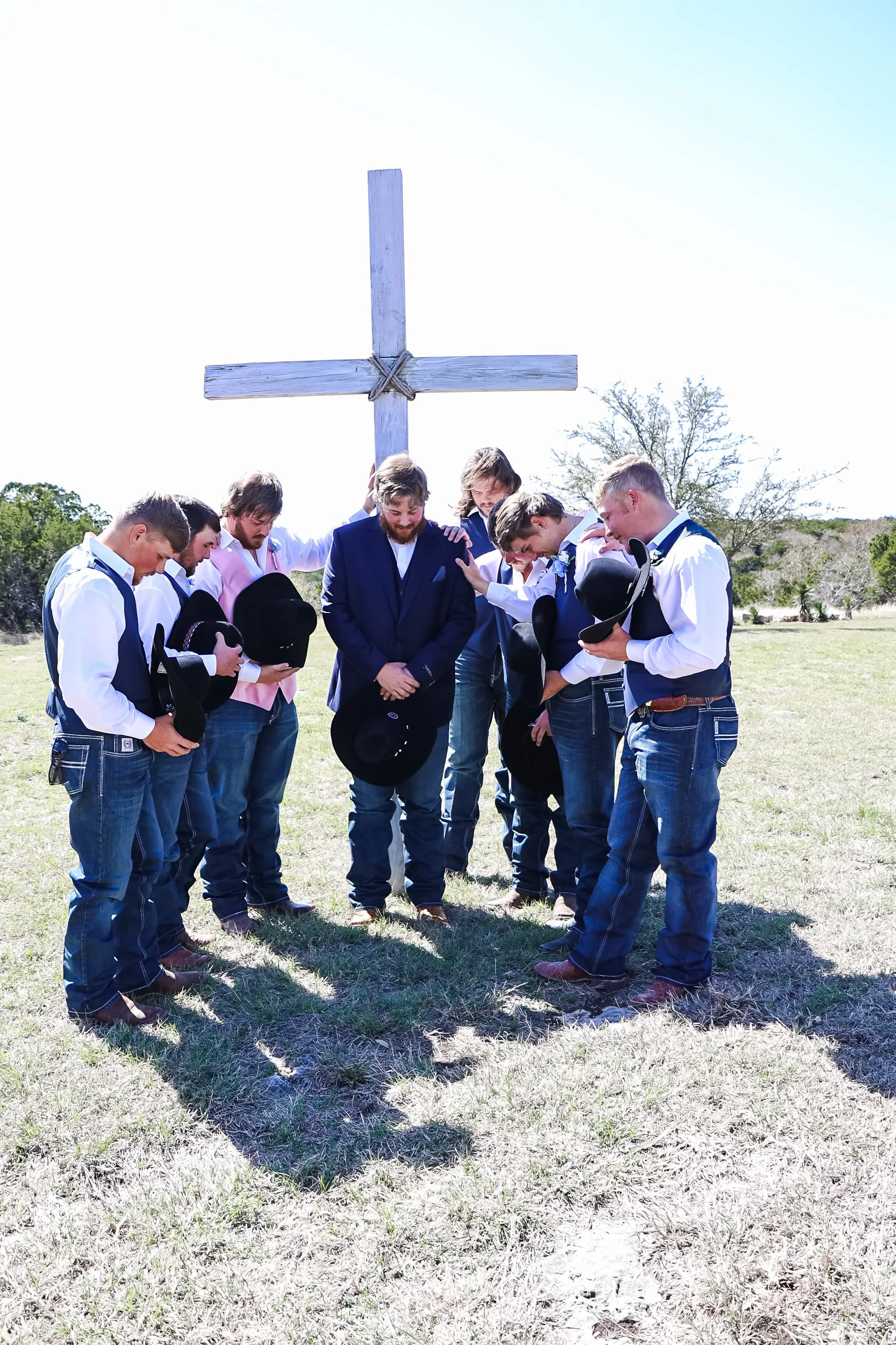 A group of men dressed in formal attire are standing in a circle outdoors, bowing their heads in prayer under a large wooden cross before the wedding in Glen Rose, Texas.