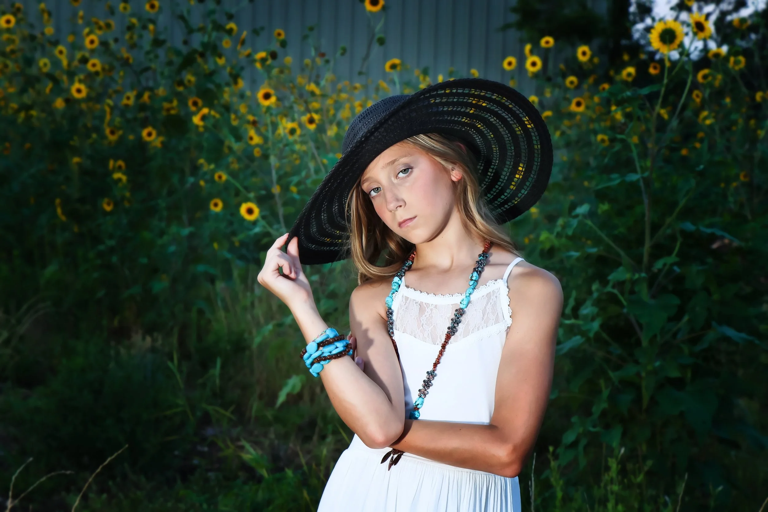 A young woman wearing a white dress and large black sun hat, standing outdoors among sunflowers, holding her hat and looking at the camera in Belton, Texas.