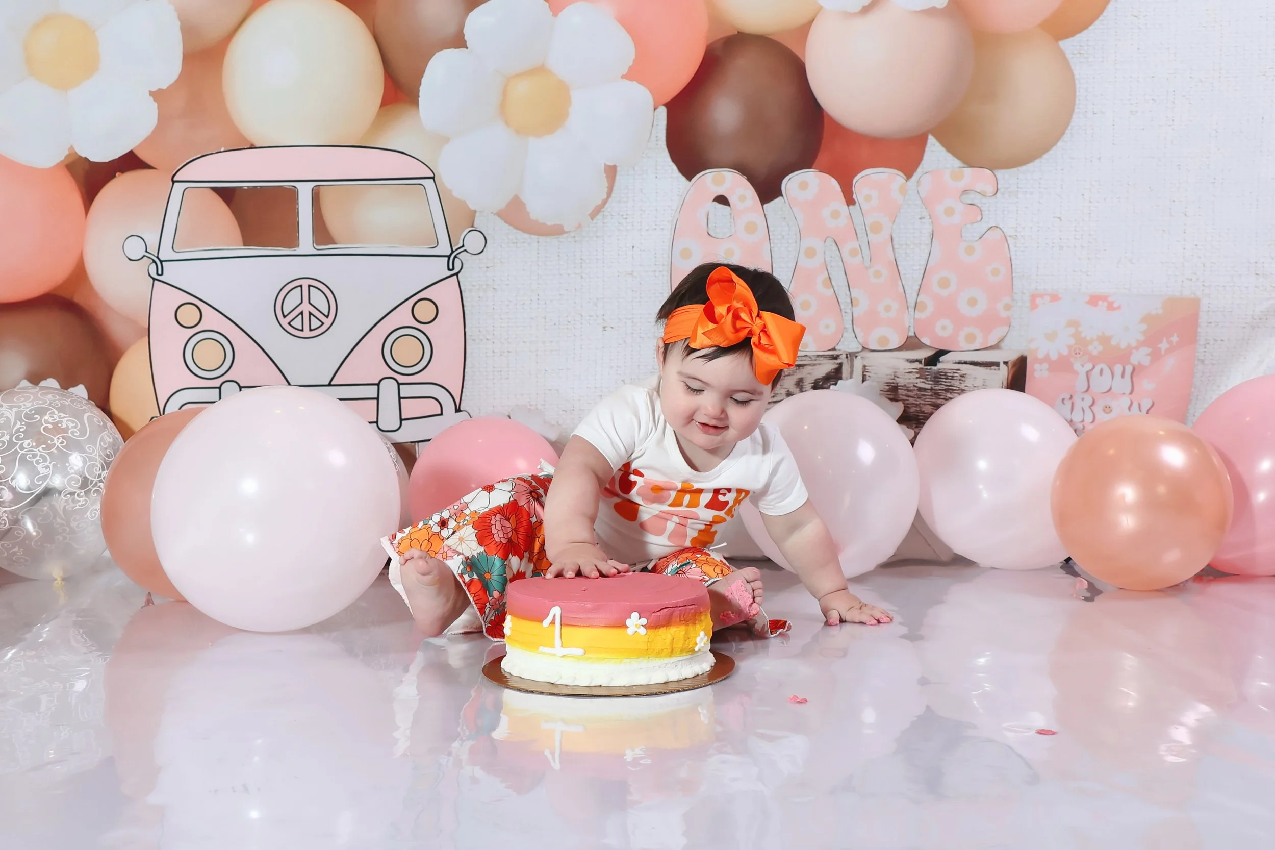 A young girl celebrating her first birthday, sitting on the floor in front of a birthday cake decorated with pink, yellow, and white layers, surrounded by pink, white, and peach balloons, with a backdrop featuring a pink and white 'LOVE' sign, a cartoon drawing of a pink and white camper van, and other decorative elements.